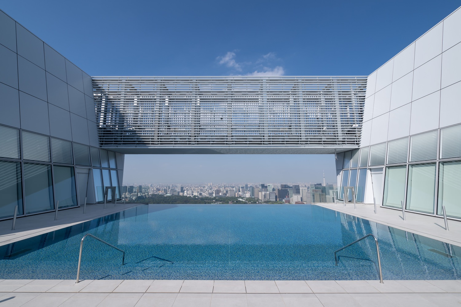 Rooftop pool terrace with two tower volumes connected by a horizontal screened bridge under blue sky