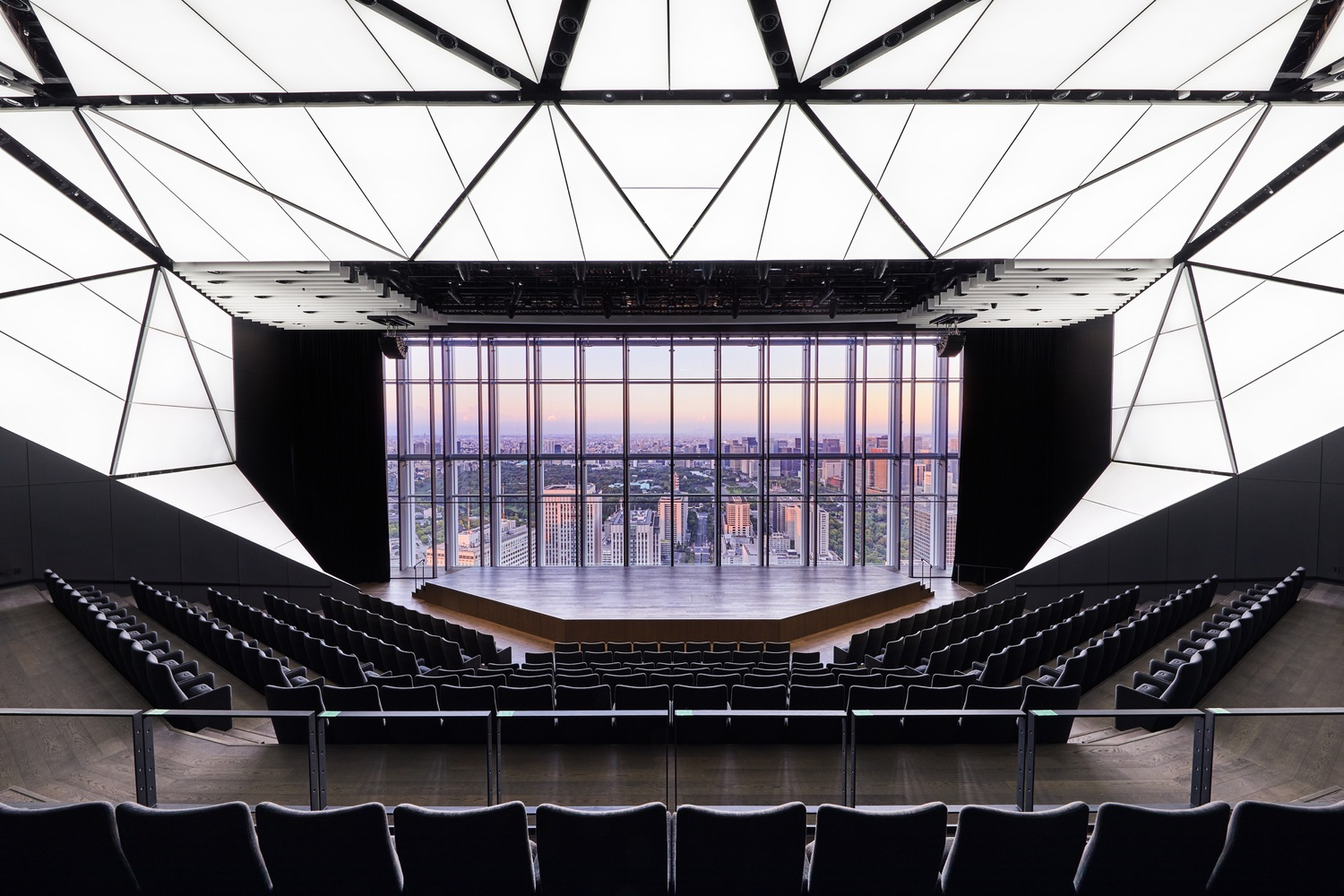 Theater interior with tiered seating facing a stage framed by floor-to-ceiling glazing at dusk