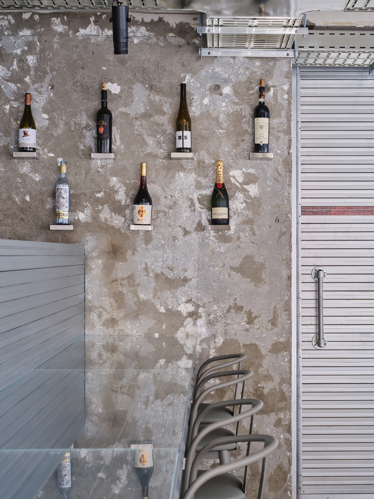 Weathered concrete wall displaying wine bottles on mounted shelves with stacked chairs below