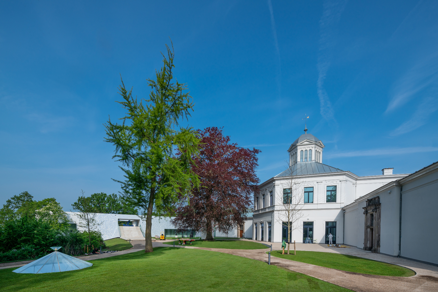 Lawn connecting white dome-topped building to low white wing with trees under clear blue sky