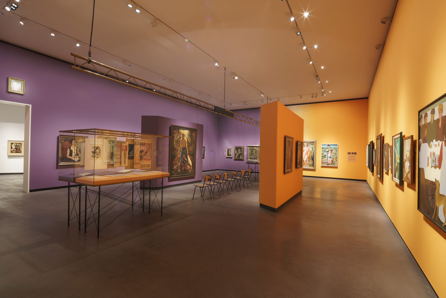 Gallery interior with purple and orange walls displaying framed artworks and a central vitrine