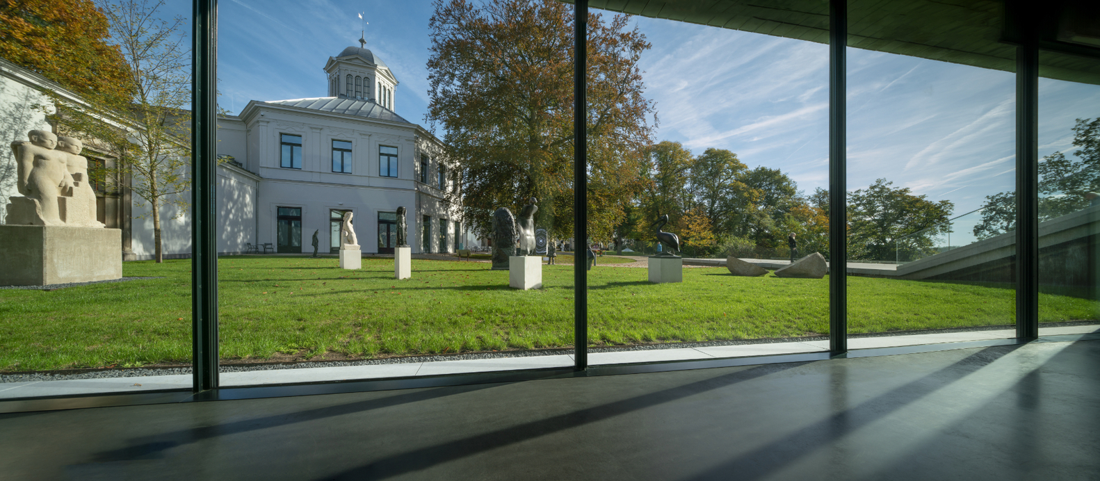 Floor-to-ceiling glass wall framing view of domed building and sculptures on lawn in autumn