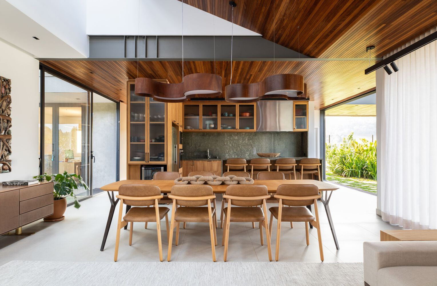 Open-plan dining area with timber ceiling and overhead pendant lights connecting to kitchen and garden through glazing