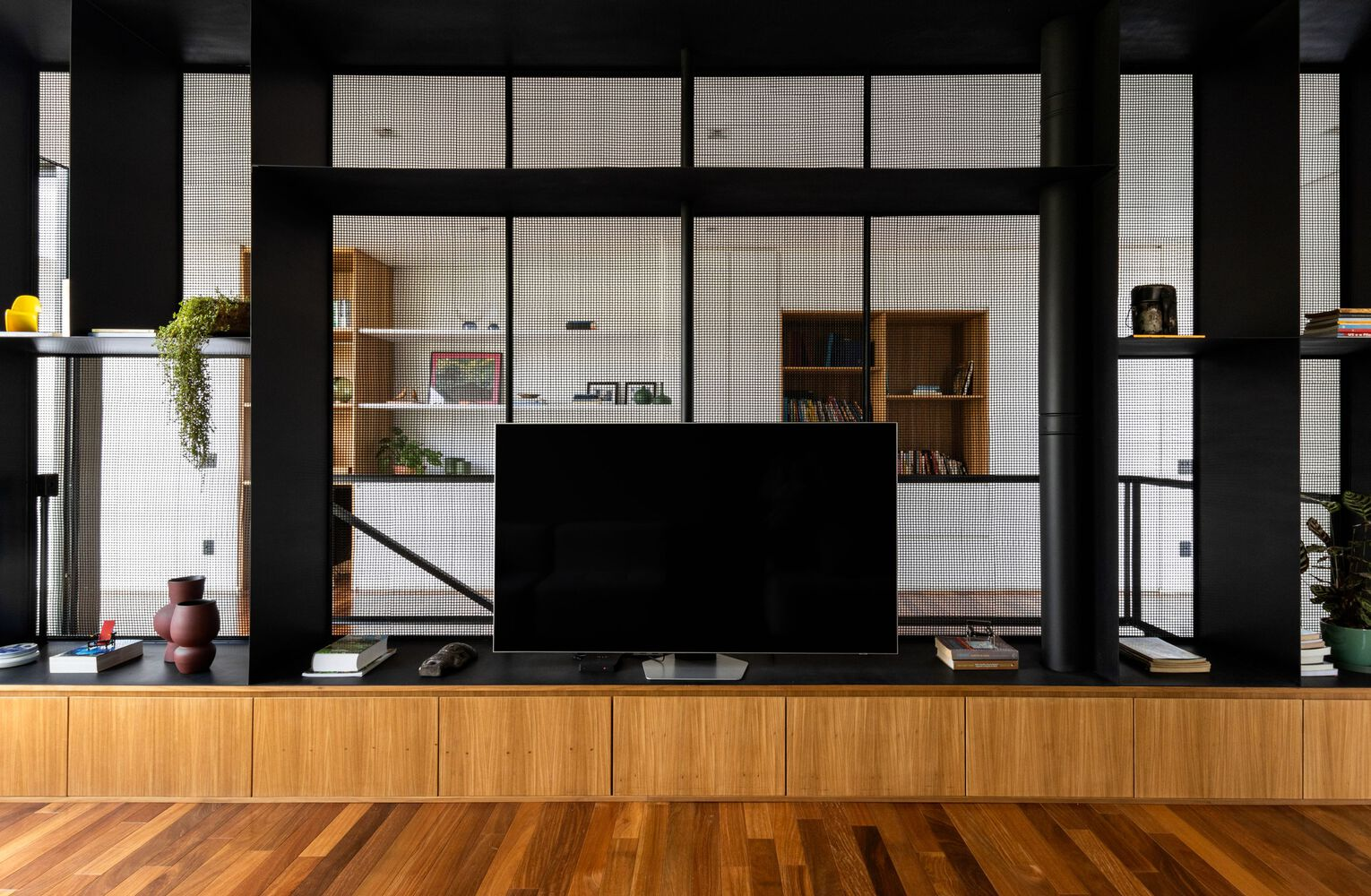 Timber credenza beneath flat-screen television framed by black steel gridded partition with integrated shelving beyond