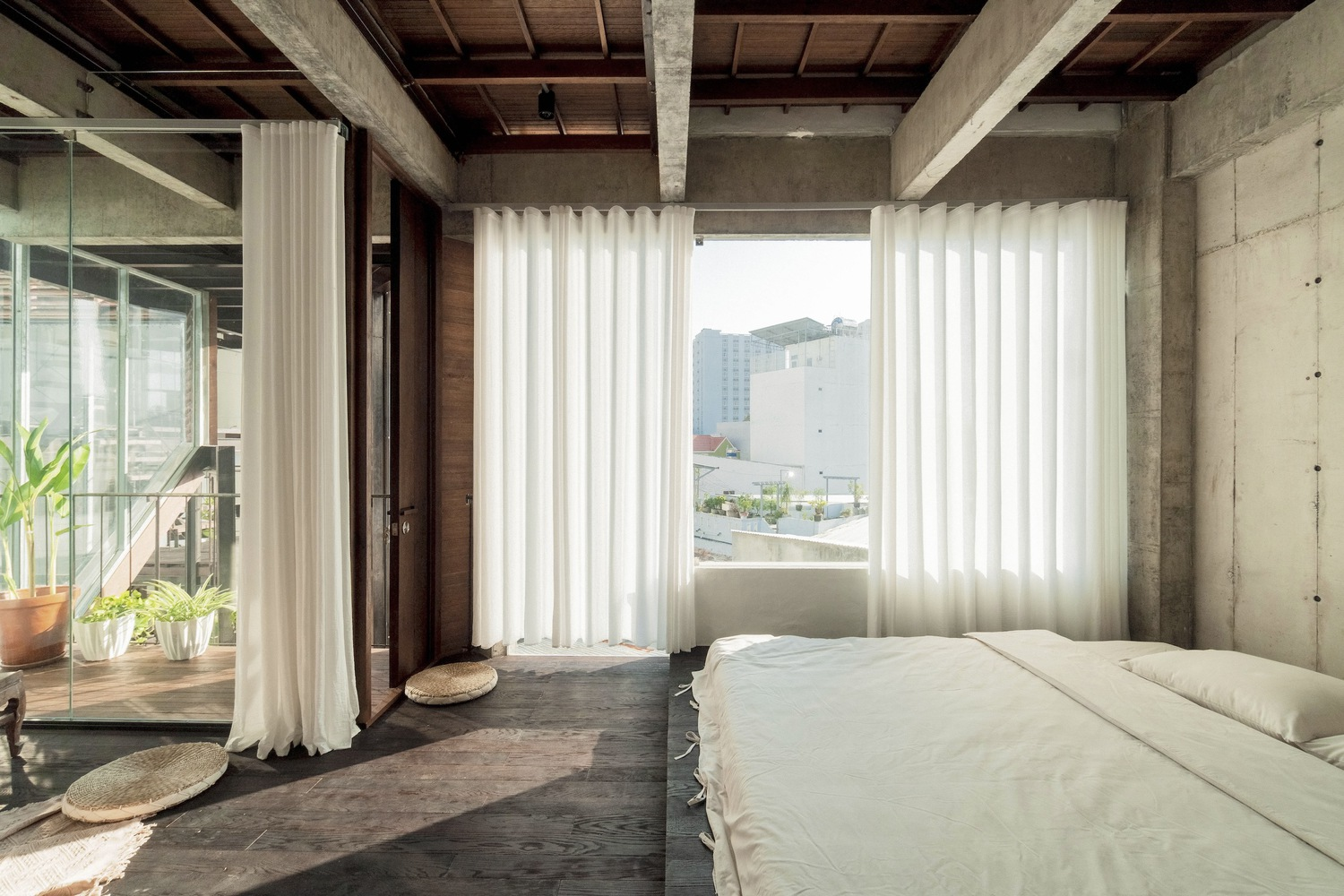 Bedroom with exposed timber ceiling, concrete columns, and sheer curtains framing the view to adjacent buildings