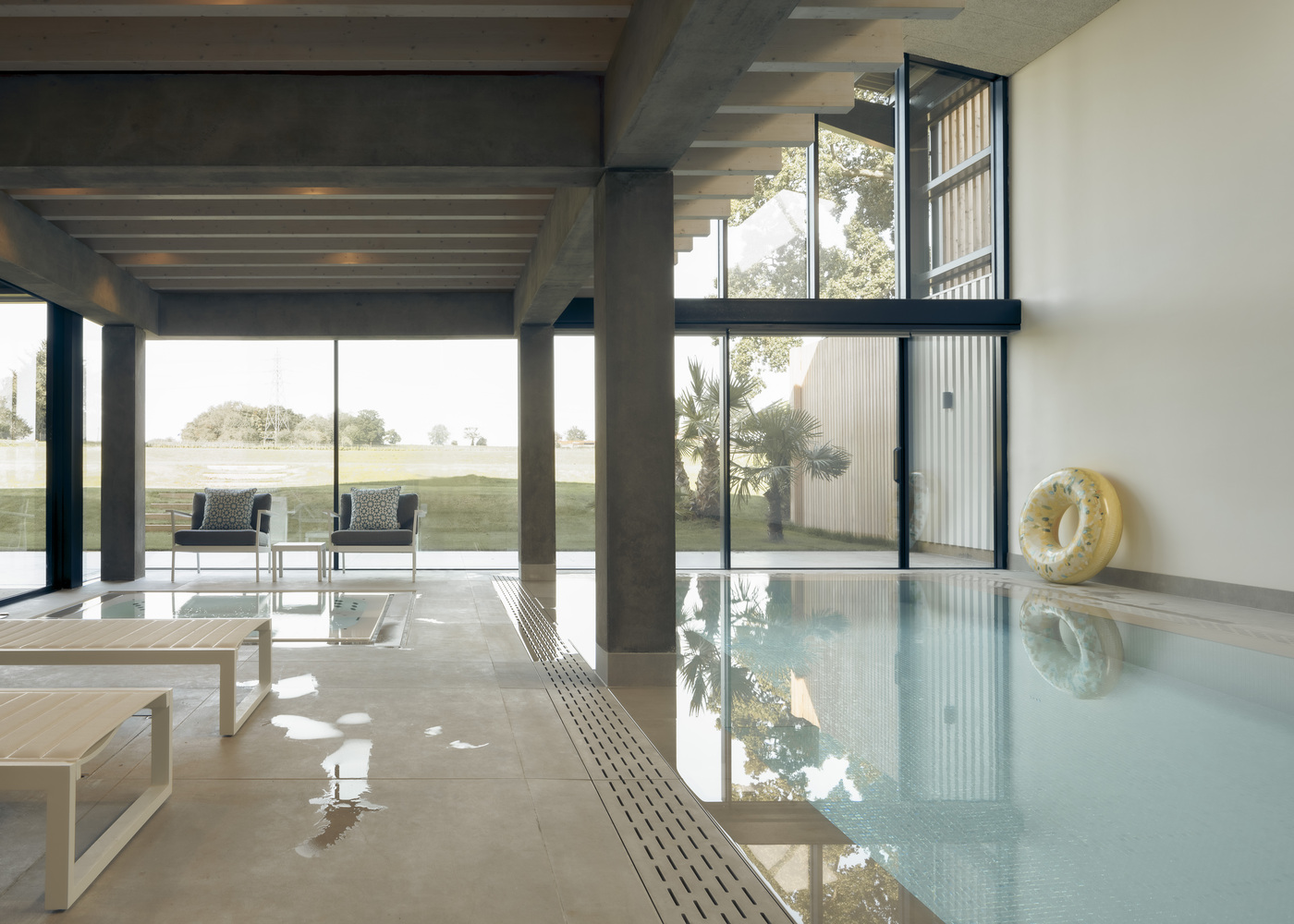 Indoor pool with tile edge and drainage channel beside glass walls opening to landscape with palms