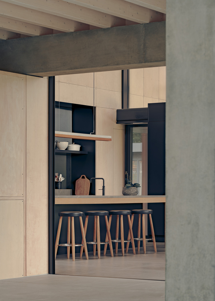 Kitchen with timber bar stools and pale plywood cabinetry framed by concrete column