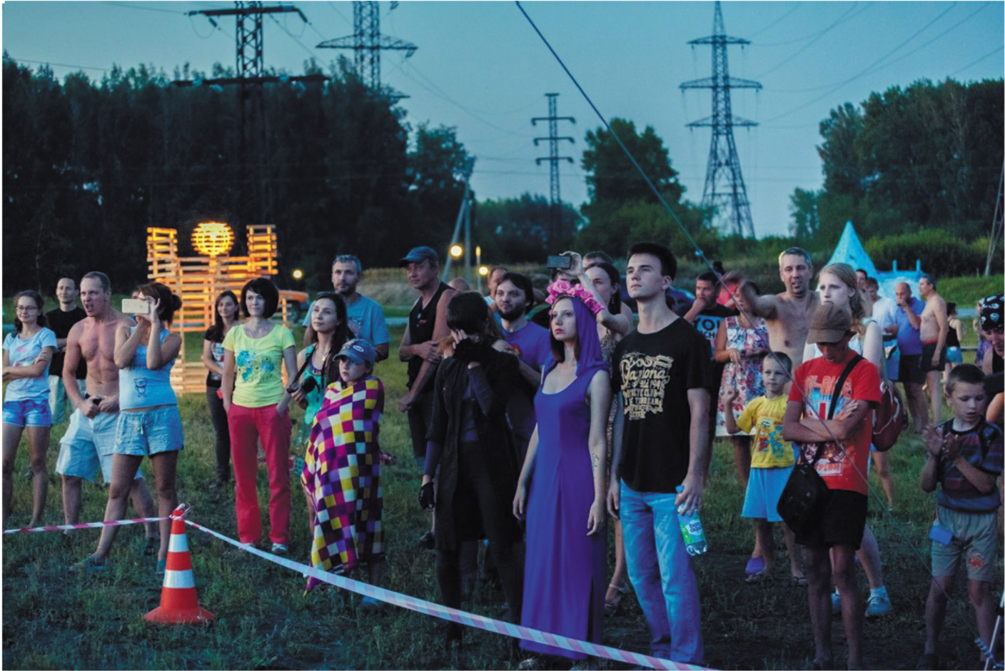Crowd gathered at dusk near transmission towers and a glowing timber installation in an open field