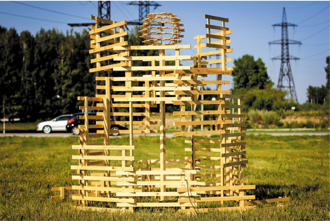 Stacked timber slat pavilion with spherical core standing in a grass field beside a roadway at sunset