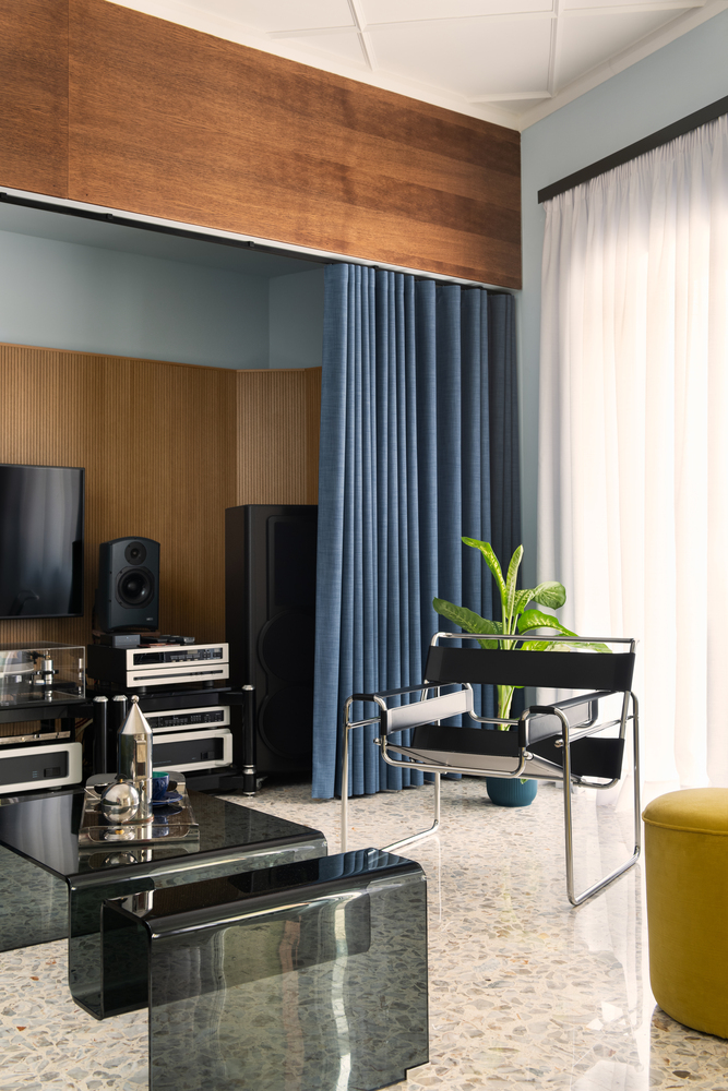 Timber-clad media alcove with blue drapery and chrome-framed lounge chair beside a glazed low table
