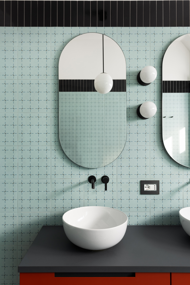 Oval mirror above a pedestal sink on a dark countertop against mint tile with black accent band