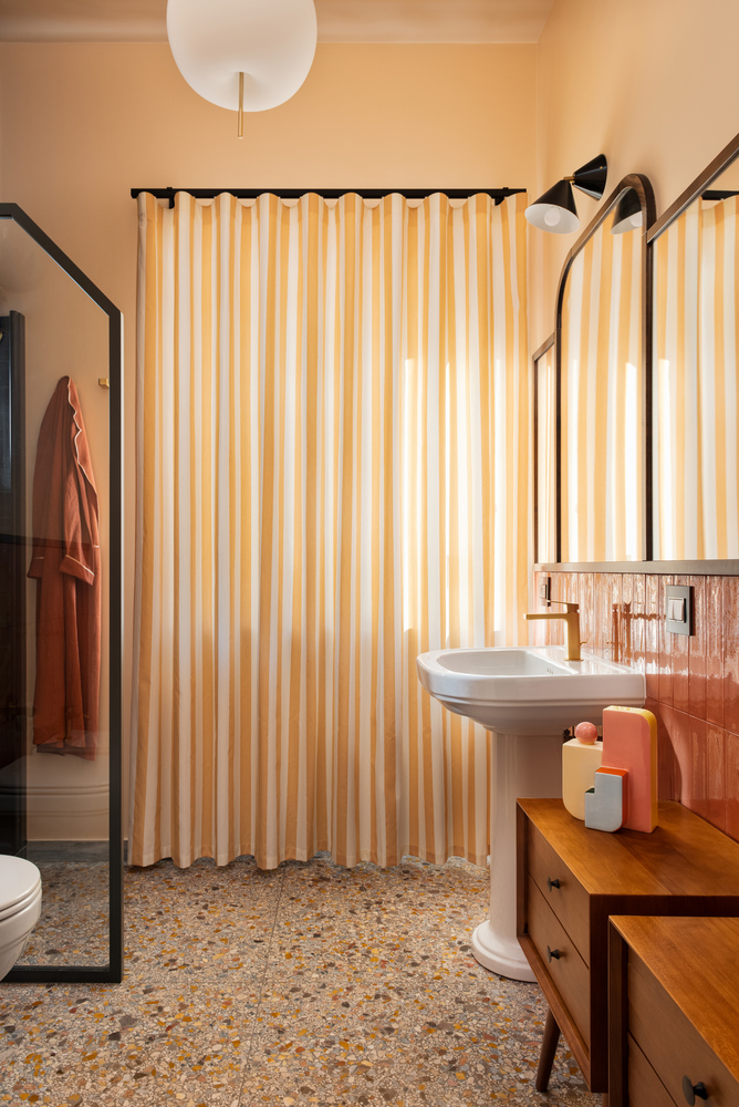 Bathroom with vertical striped curtain, terrazzo floor, timber vanity, and arched mirror in warm light