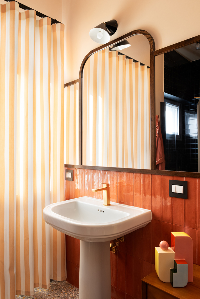 Wall-mounted pedestal sink below arched mirror with orange tile backsplash and vertical striped wallpaper