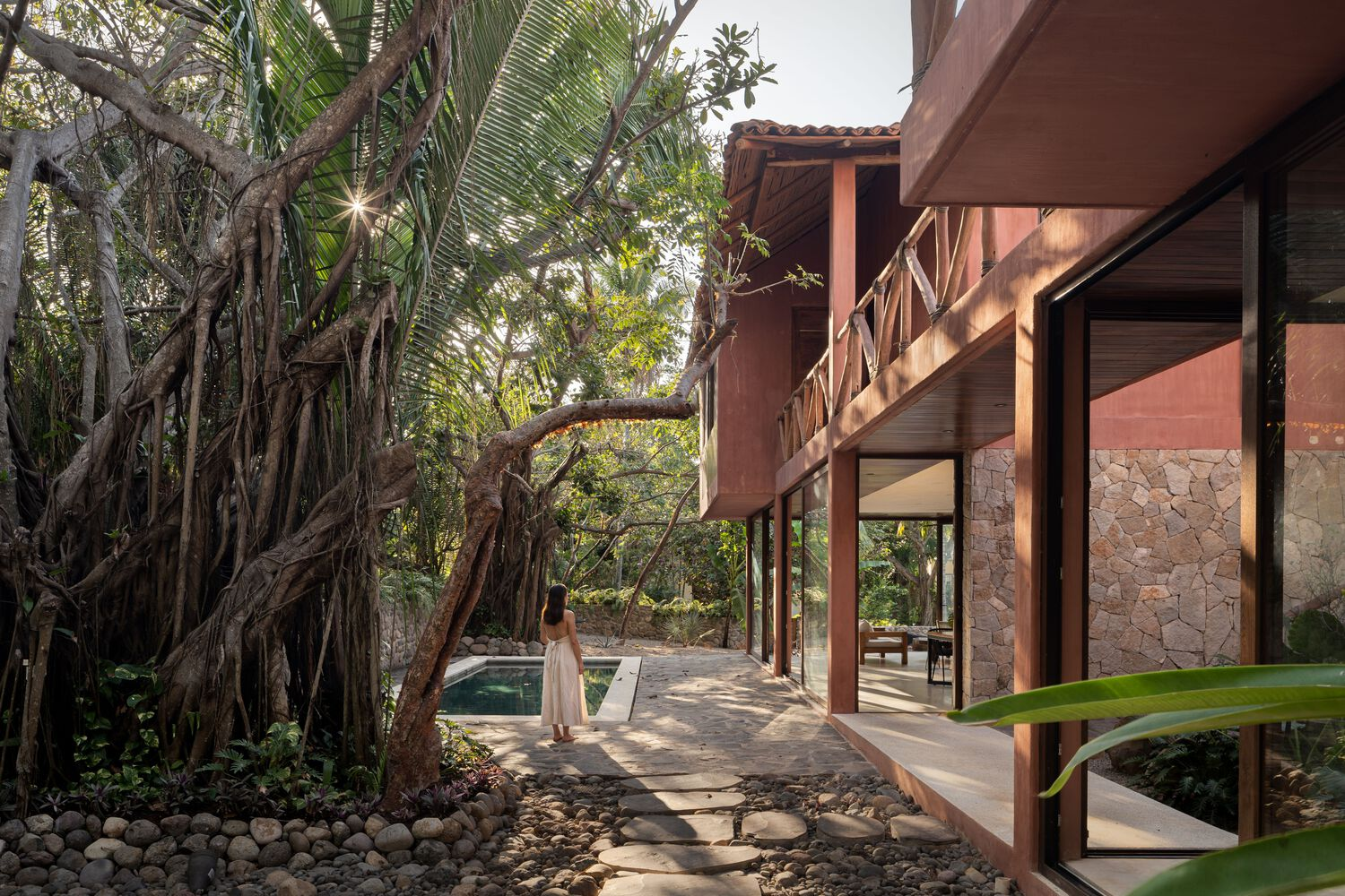Covered walkway with timber columns and stone walls alongside a pool and banyan tree roots