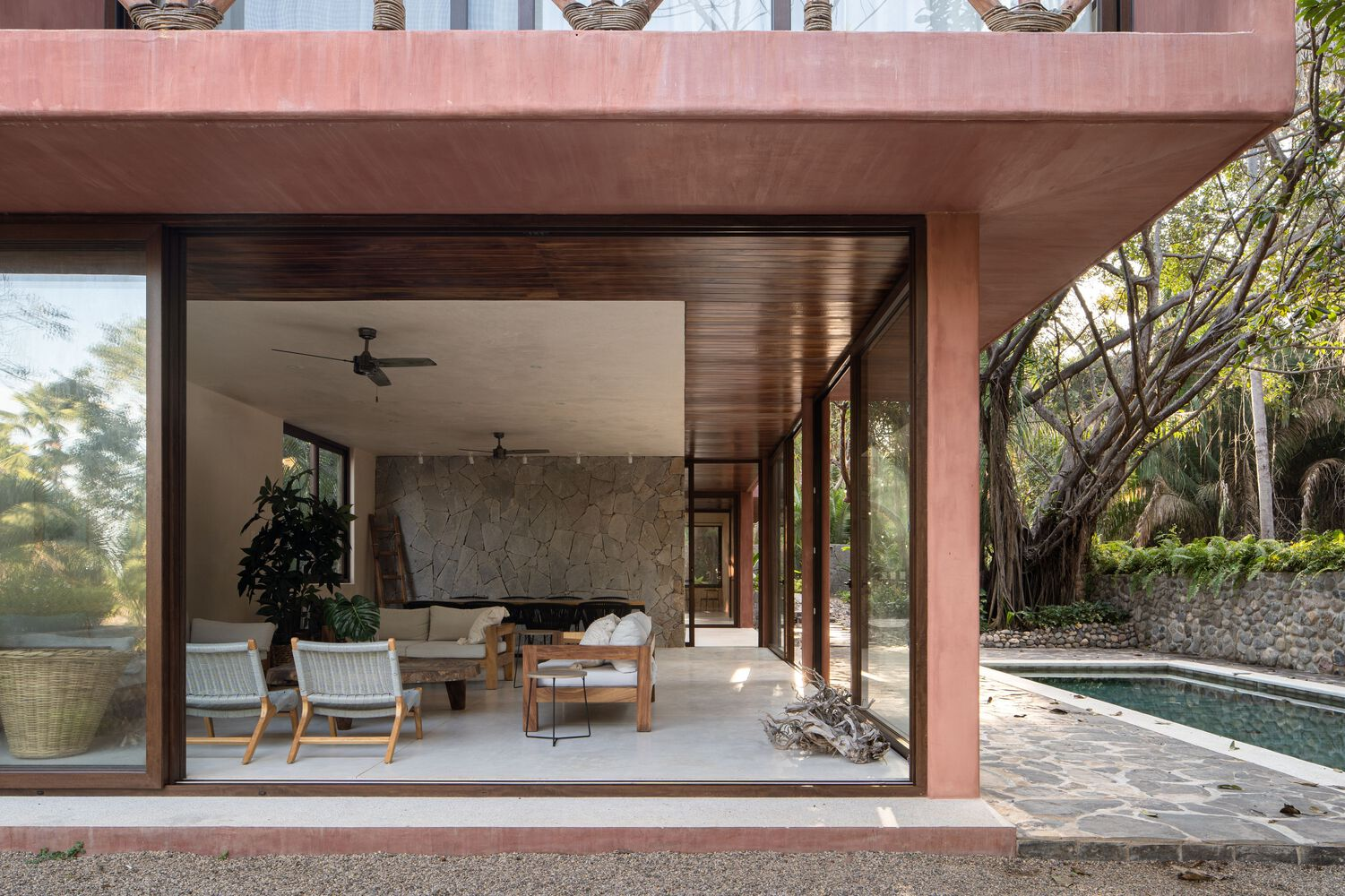 Living room with slatted timber ceiling and stone wall opening to poolside terrace in afternoon light