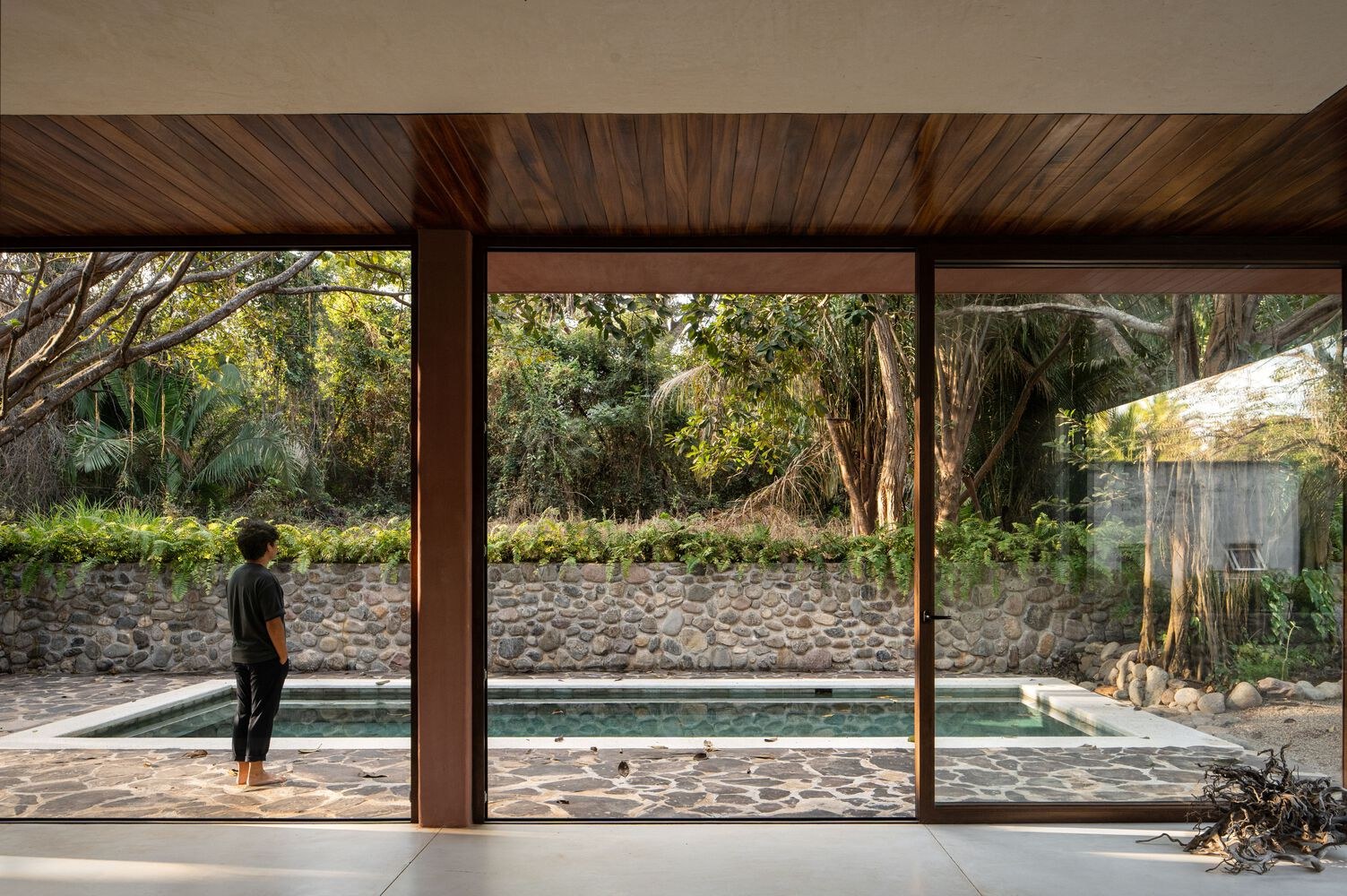 Floor-to-ceiling glass wall framing the pool and cobblestone retaining wall with a figure standing inside