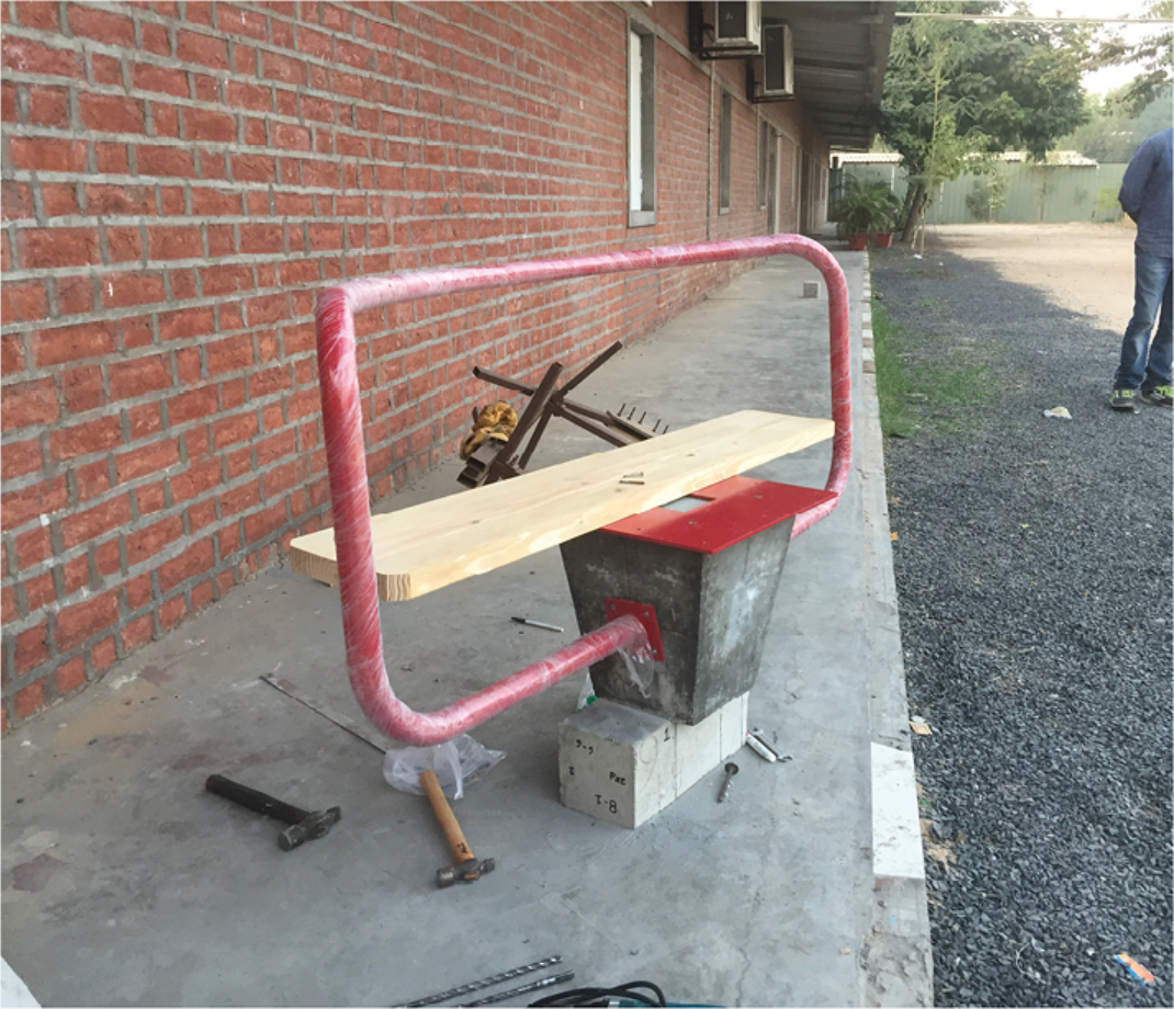 Prototype bench under construction with pink-wrapped steel frame and timber plank beside a brick wall