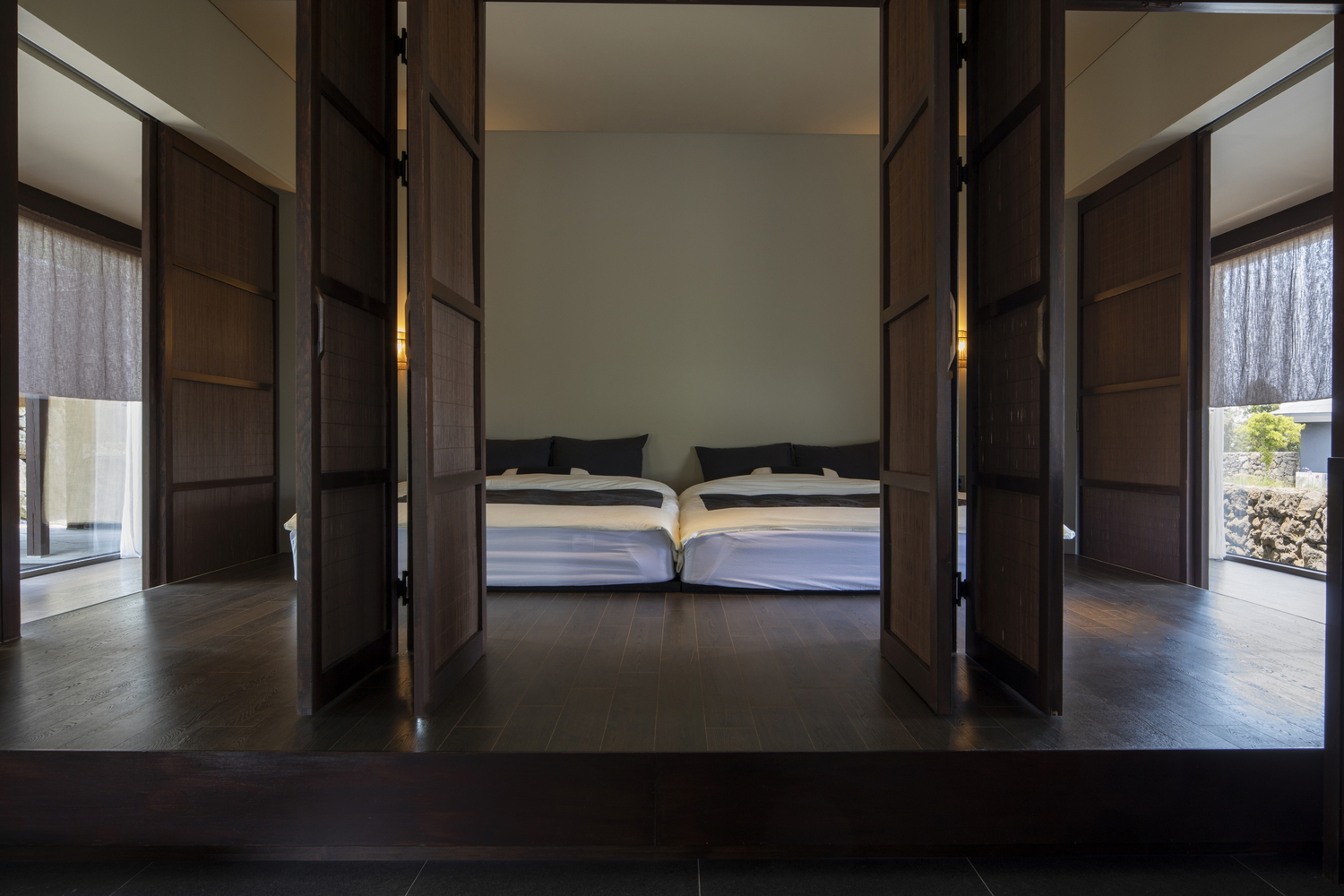 View through open dark timber folding doors into bedroom with twin beds and wooden flooring