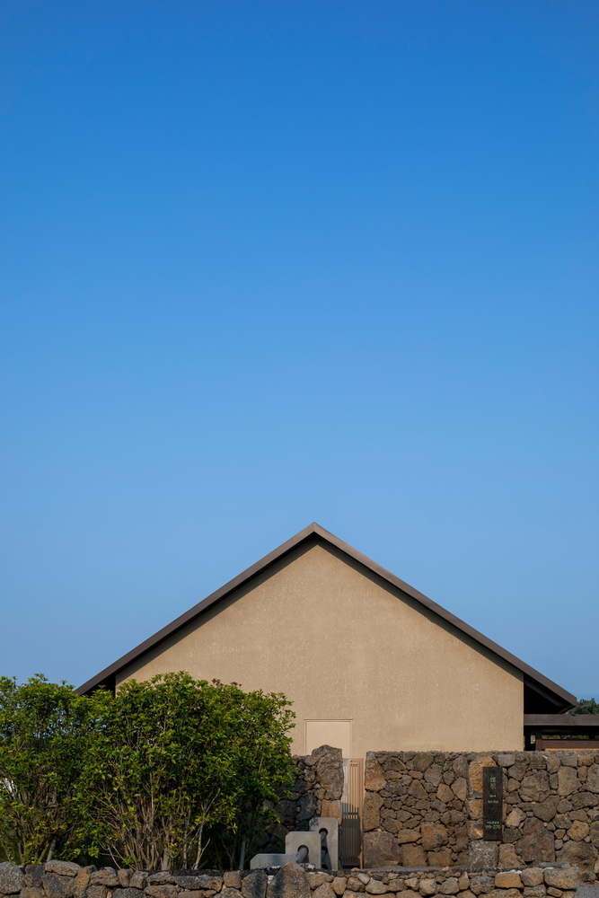 Gabled facade in sand-colored plaster rising above volcanic stone base wall with green foliage