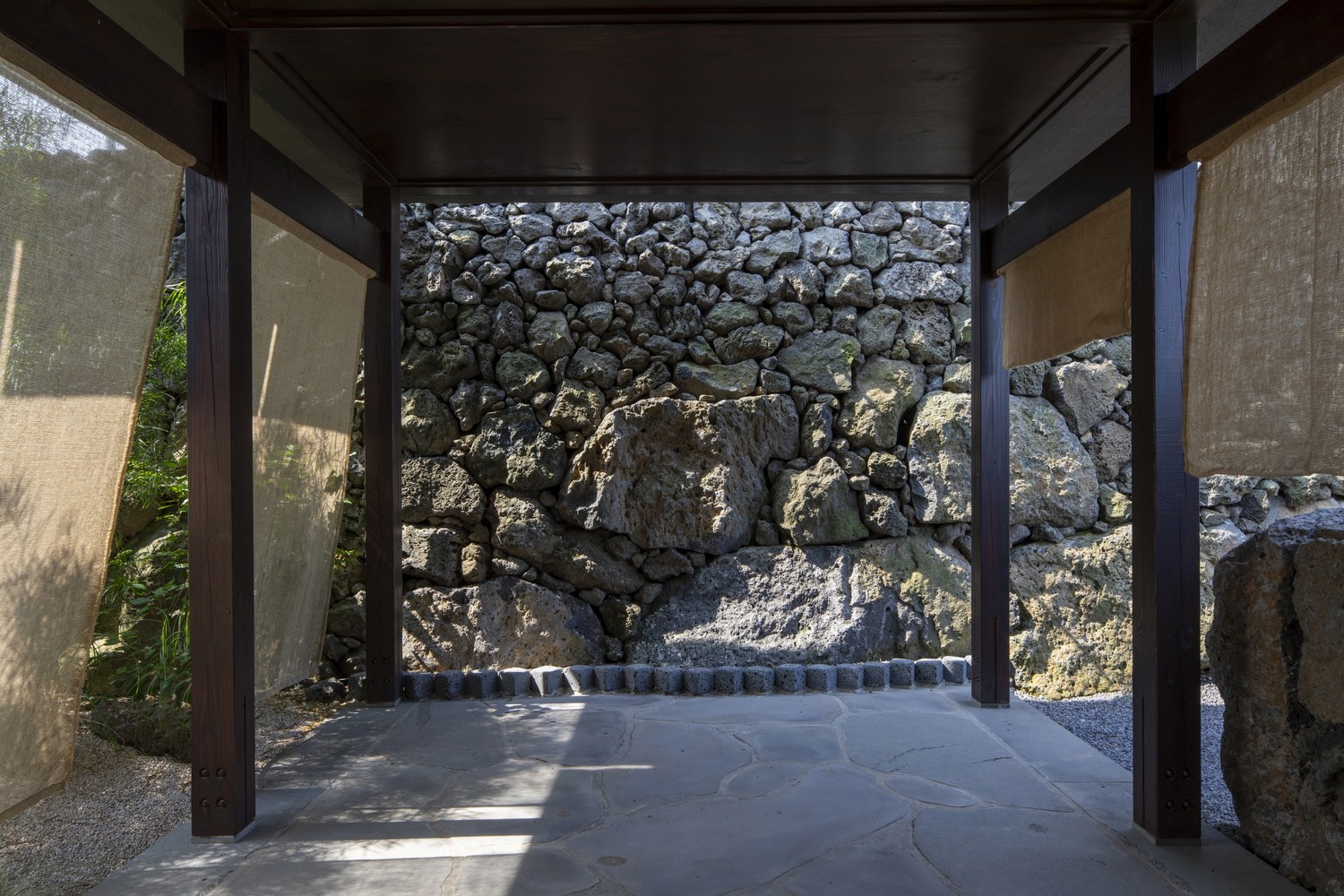 Covered passage framing rough volcanic stone wall with flag pavers and gravel in dappled sunlight