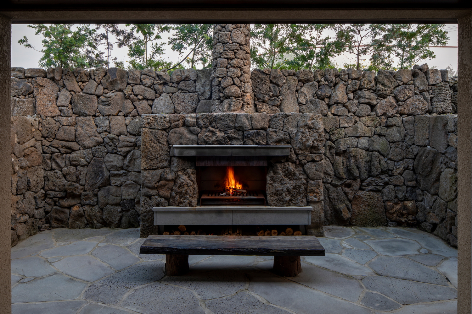 Outdoor fireplace set into stacked volcanic stone wall with timber bench and flagstone terrace at dusk