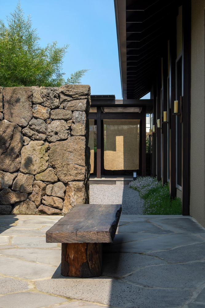 Outdoor courtyard with rough volcanic stone wall, timber bench and steel-framed glazing under clear blue sky
