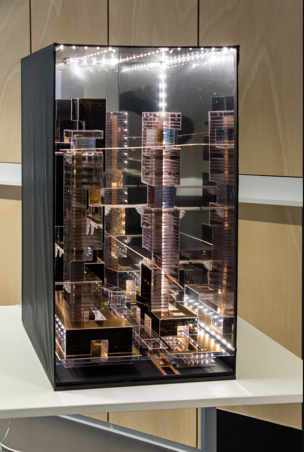 Physical model in a glass vitrine showing layered urban towers with internal LED lighting strips