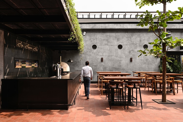 Courtyard terrace with metal bar counter, circular porthole windows in concrete wall and potted trees between tables