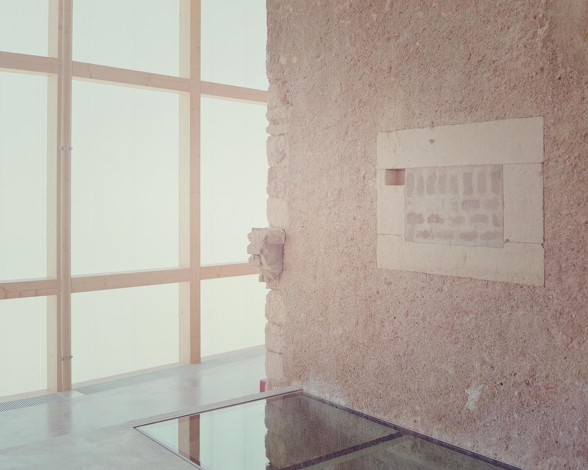 Corner detail of exposed stone wall with framed recess beside translucent glass partition and floor opening