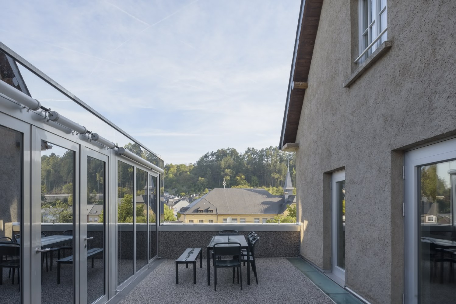 Rooftop terrace view between stucco volume and glazed pavilion with forested hillside beyond