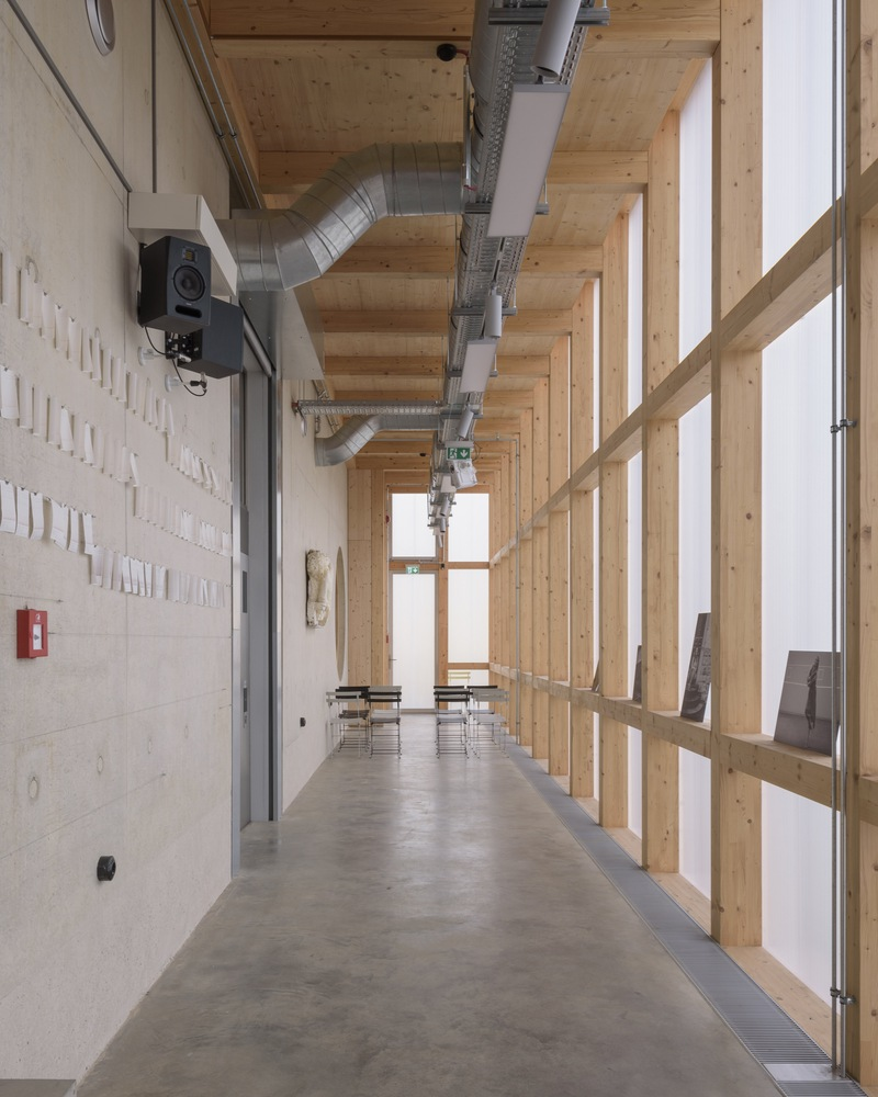 Concrete and timber corridor with exposed ventilation ducts and a diagonal timber screen along glazed facade