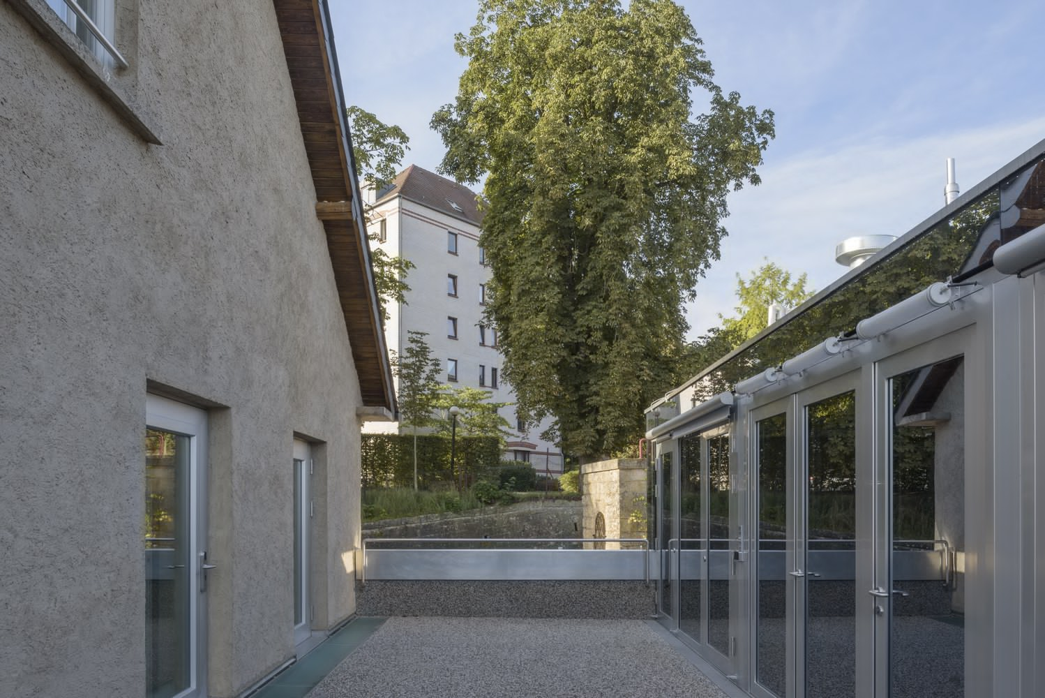 Gravel courtyard flanked by a textured stucco wall and a glazed pavilion under mature trees