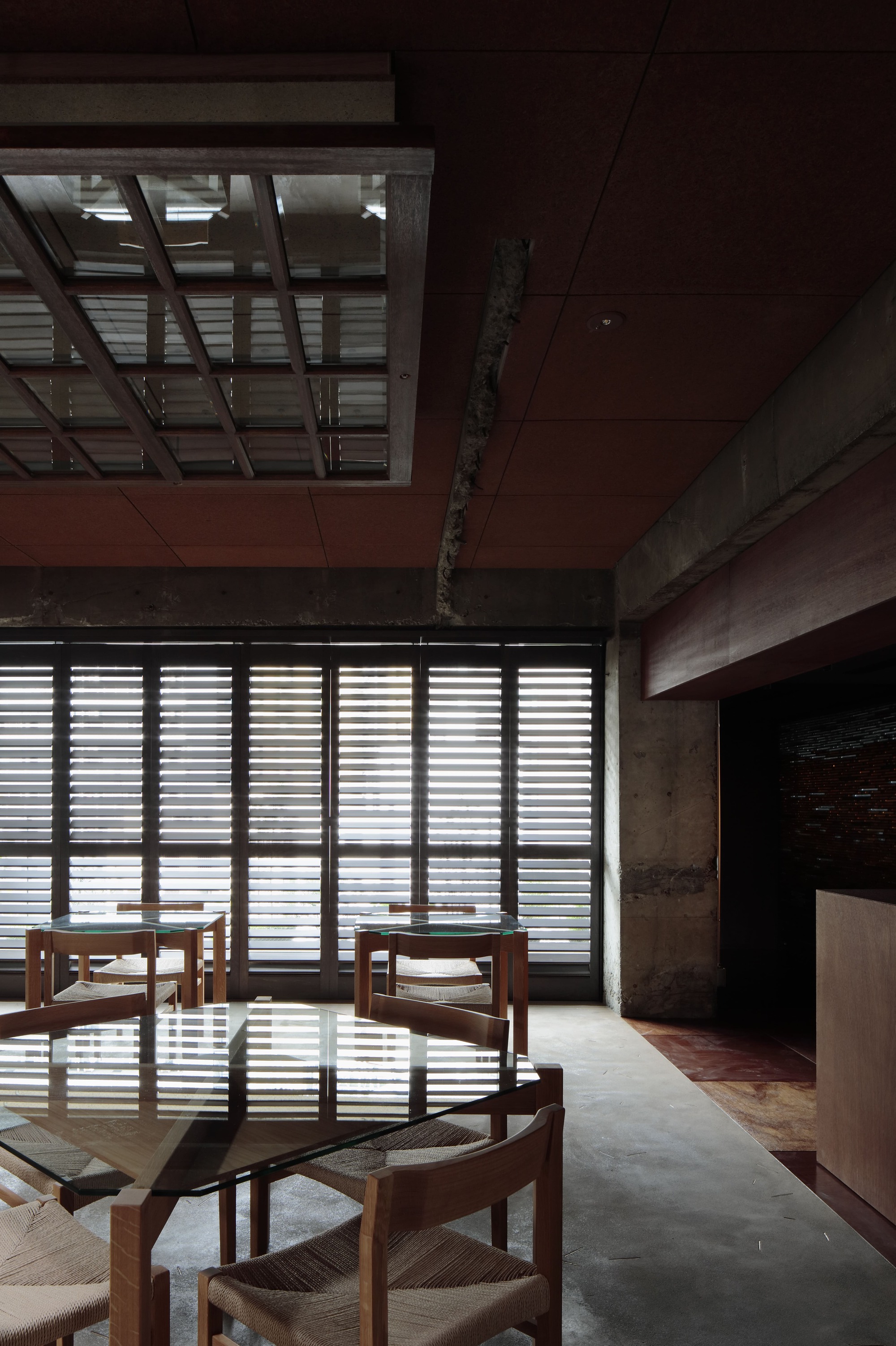 Dining chairs beside floor-to-ceiling louvered screens filtering daylight into concrete and timber interior