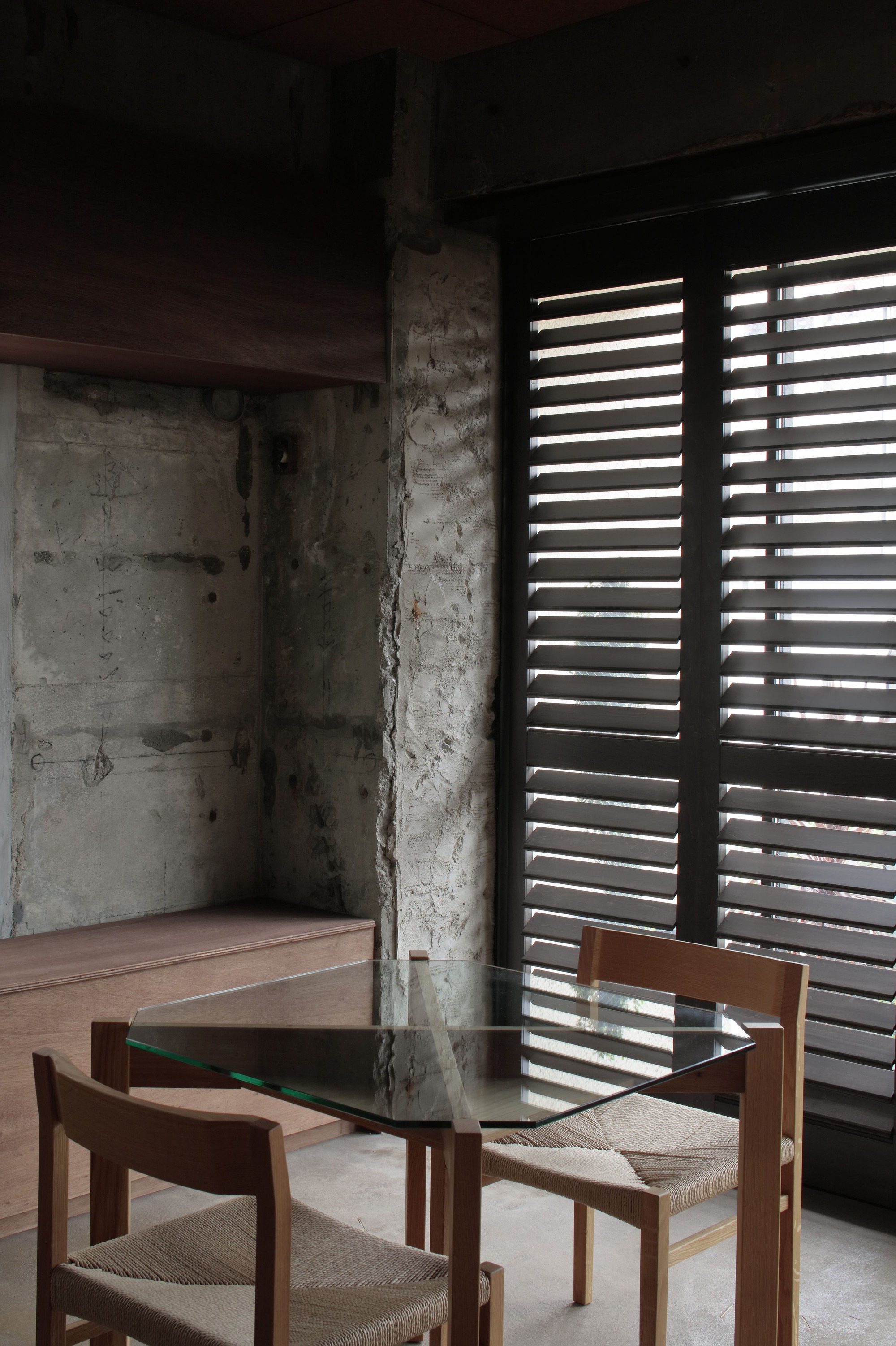 Interior corner showing exposed concrete walls with horizontal louvers filtering light beside timber and glass dining table