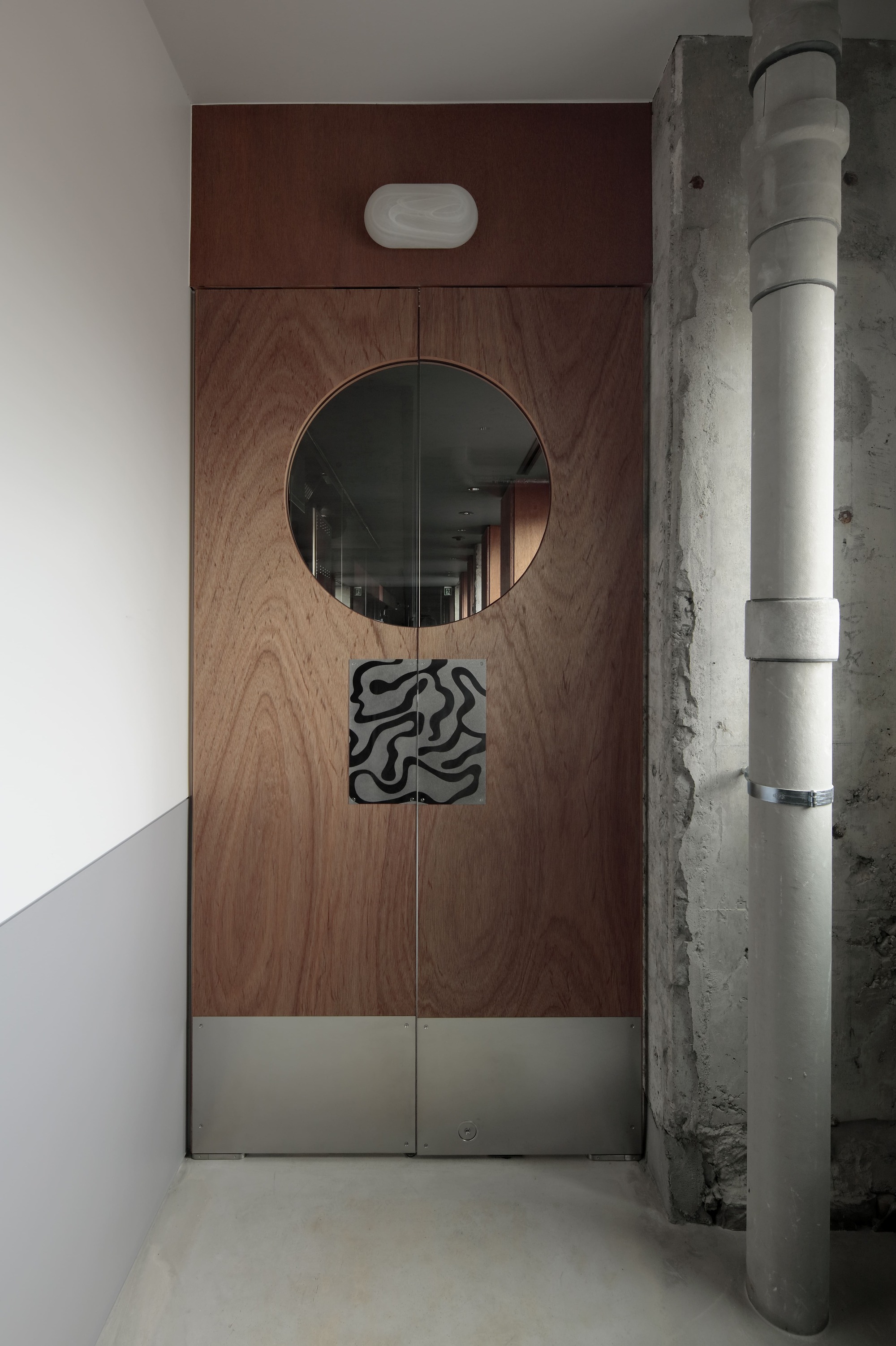 Walnut veneer entrance door with circular mirror and graphic signage beside an exposed concrete column