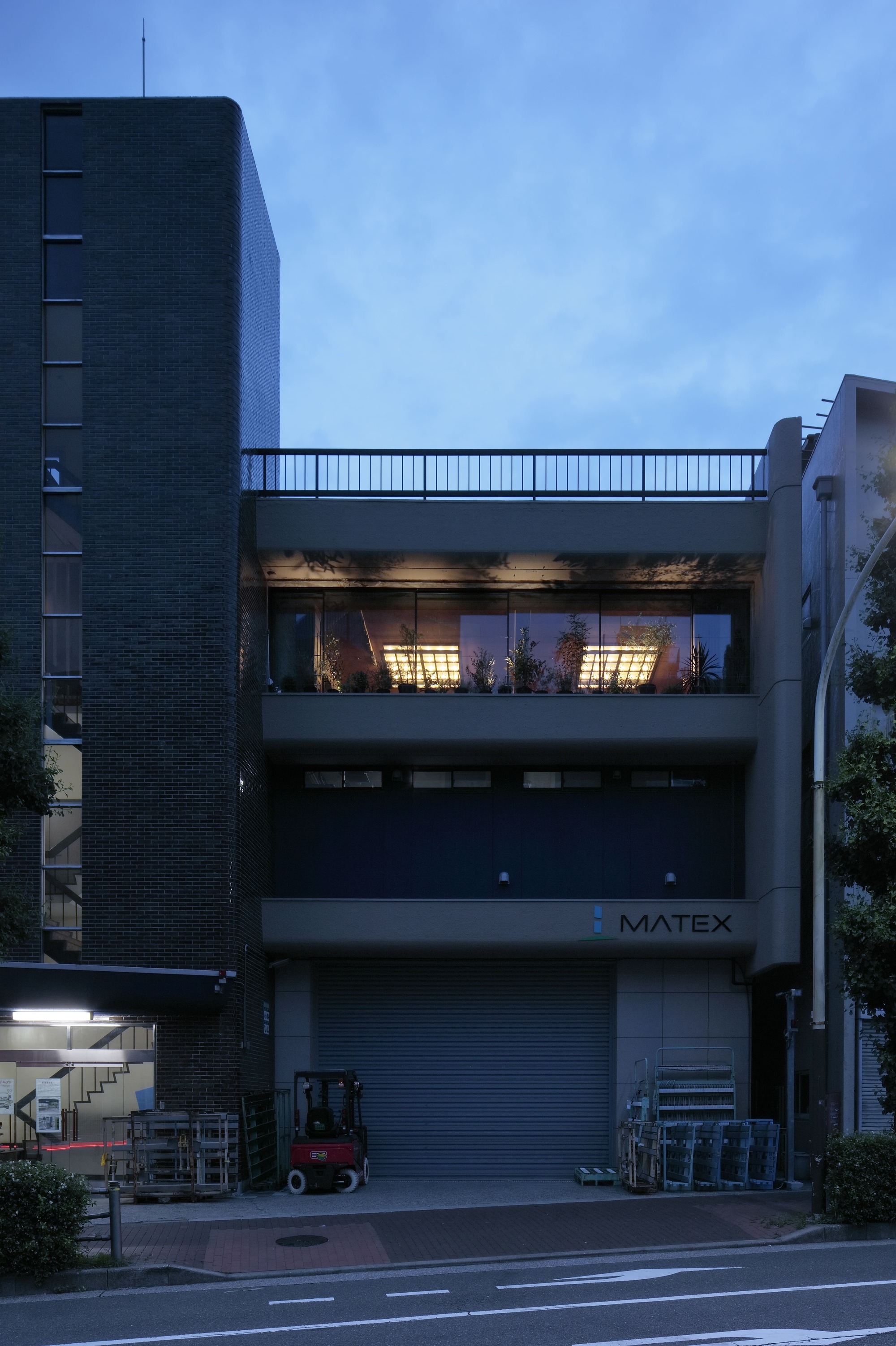 Street facade at dusk showing the second-floor terrace with illuminated interior and roll-down shutter below