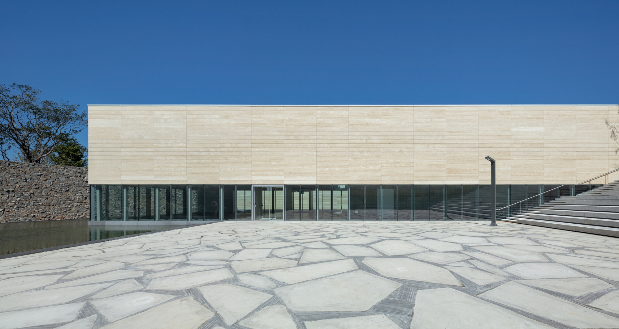 Horizontal travertine facade above a continuous glass band facing a paved stone plaza under clear blue sky