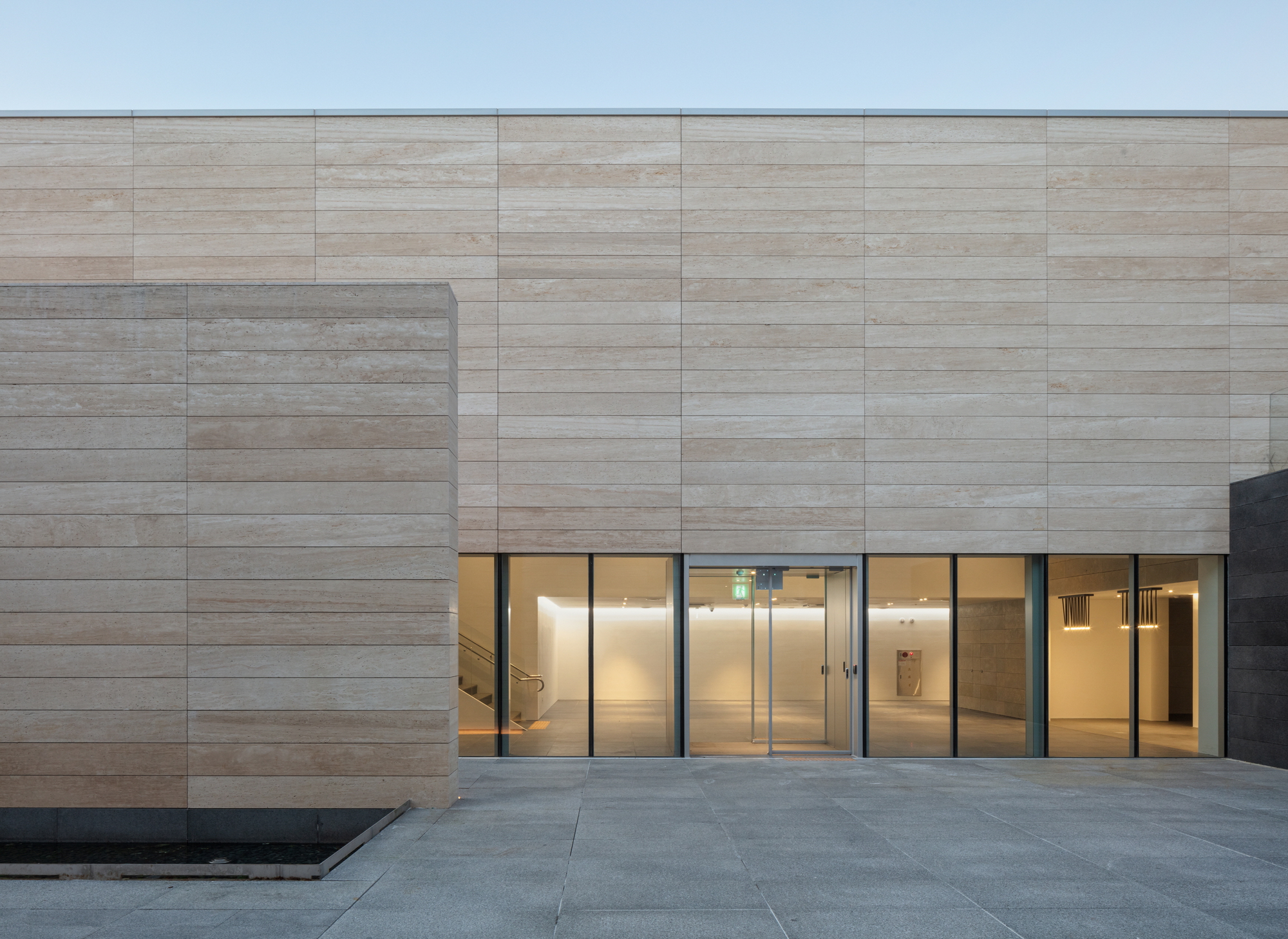 Travertine facade with recessed glass entrance and warm interior lighting at dusk