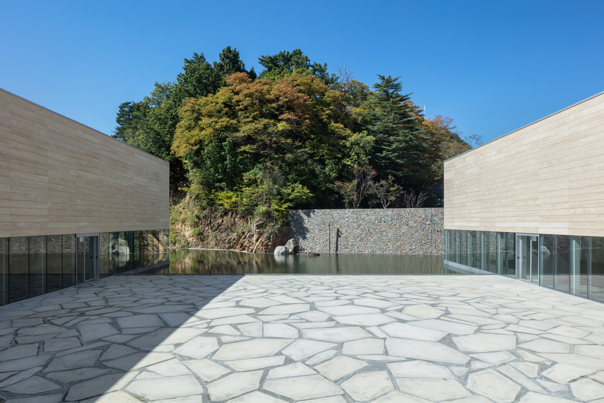 Courtyard plaza with irregular stone paving flanked by horizontal timber-clad volumes and gabion wall backdrop