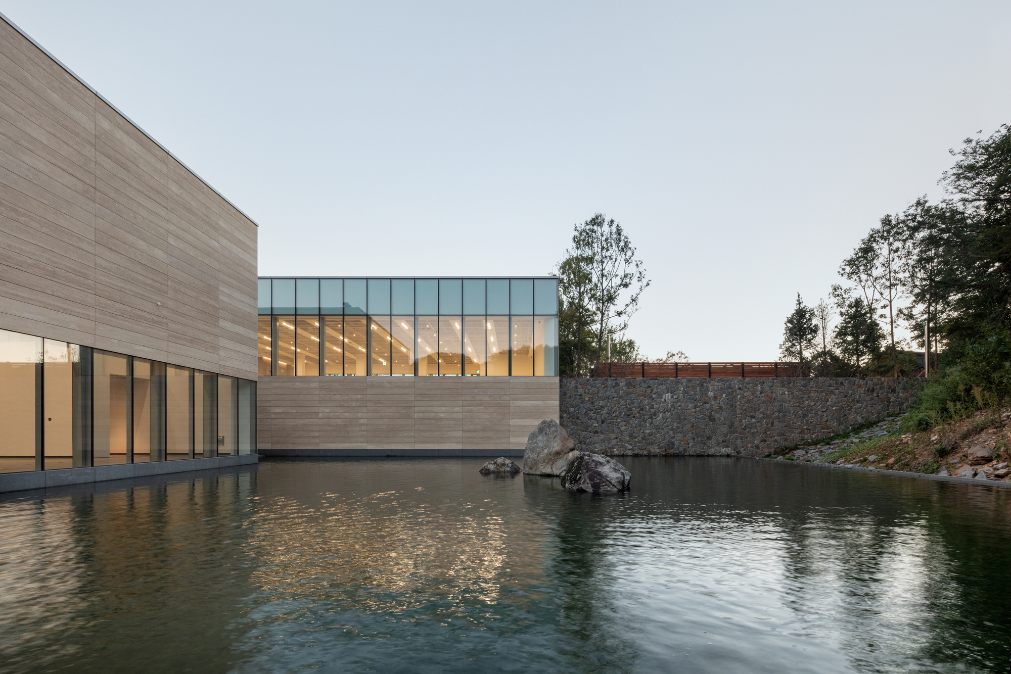 Clerestory glazing above horizontal stone-clad base beside water pool with gabion retaining wall