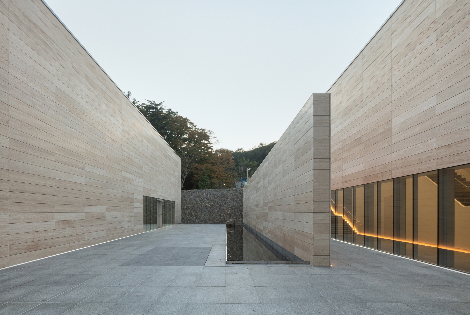 Narrow passage between parallel stone-clad walls leading to gabion-faced hillside under evening sky