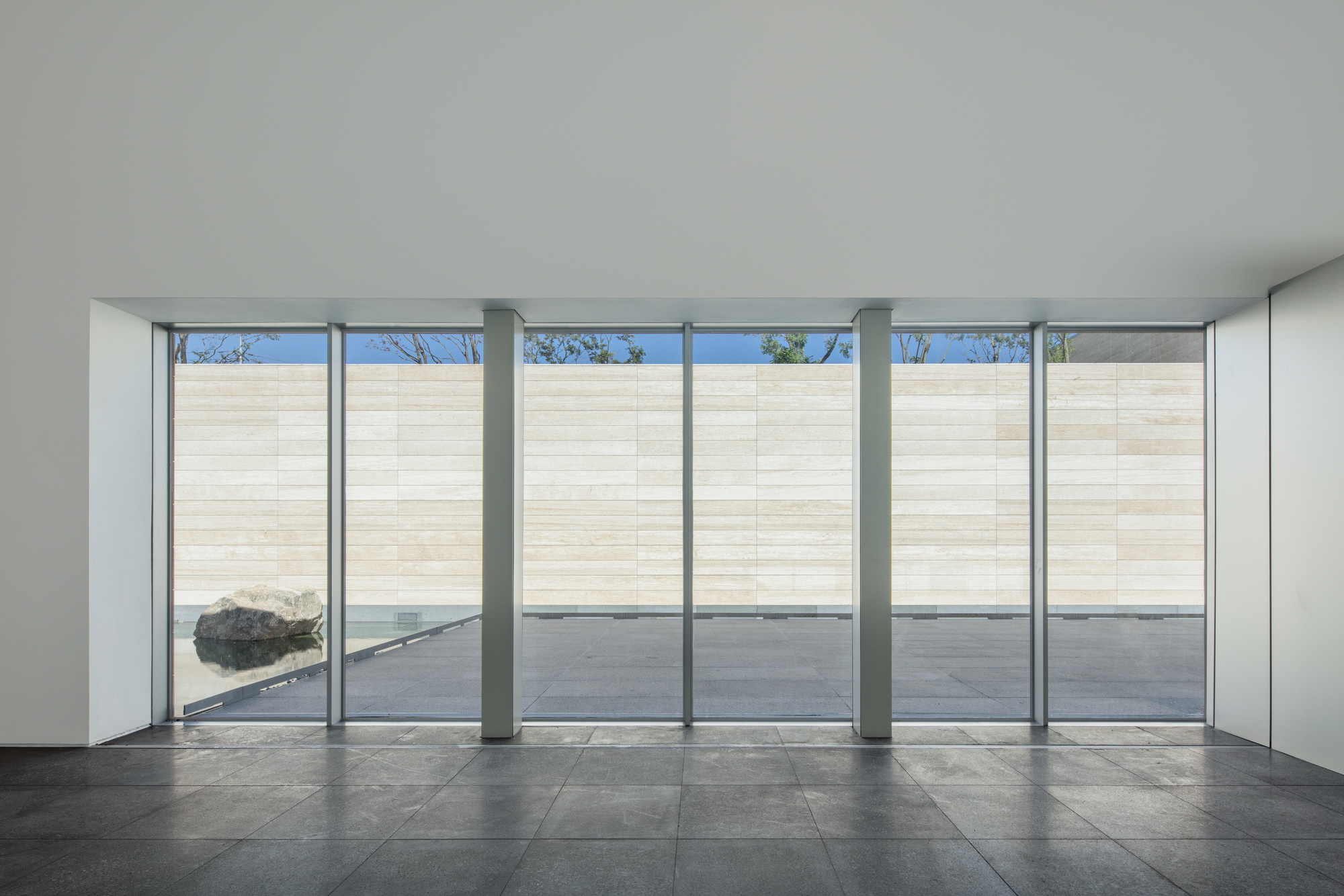Interior gallery space with floor-to-ceiling glass walls facing a pale stone courtyard wall