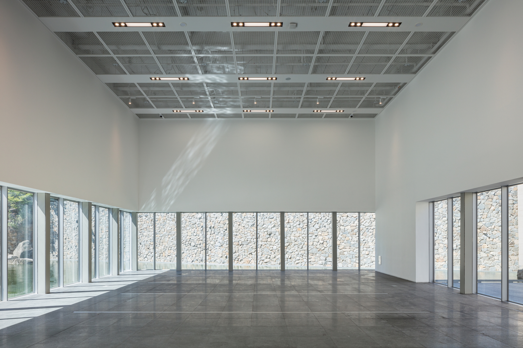 Large exhibition hall with perforated metal ceiling panels and continuous glazing overlooking a stone wall