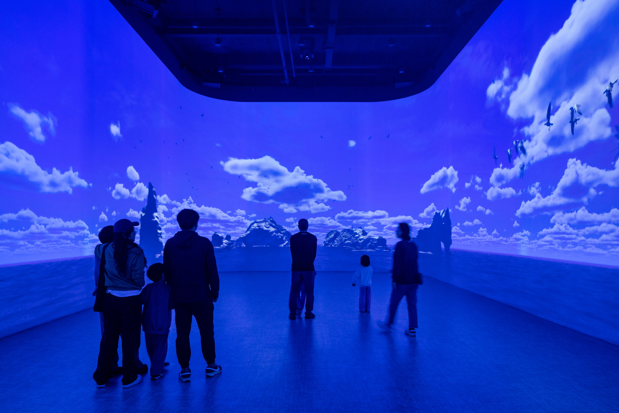 Immersive projection room bathed in blue light showing mountain and cloud imagery with visitors silhouetted