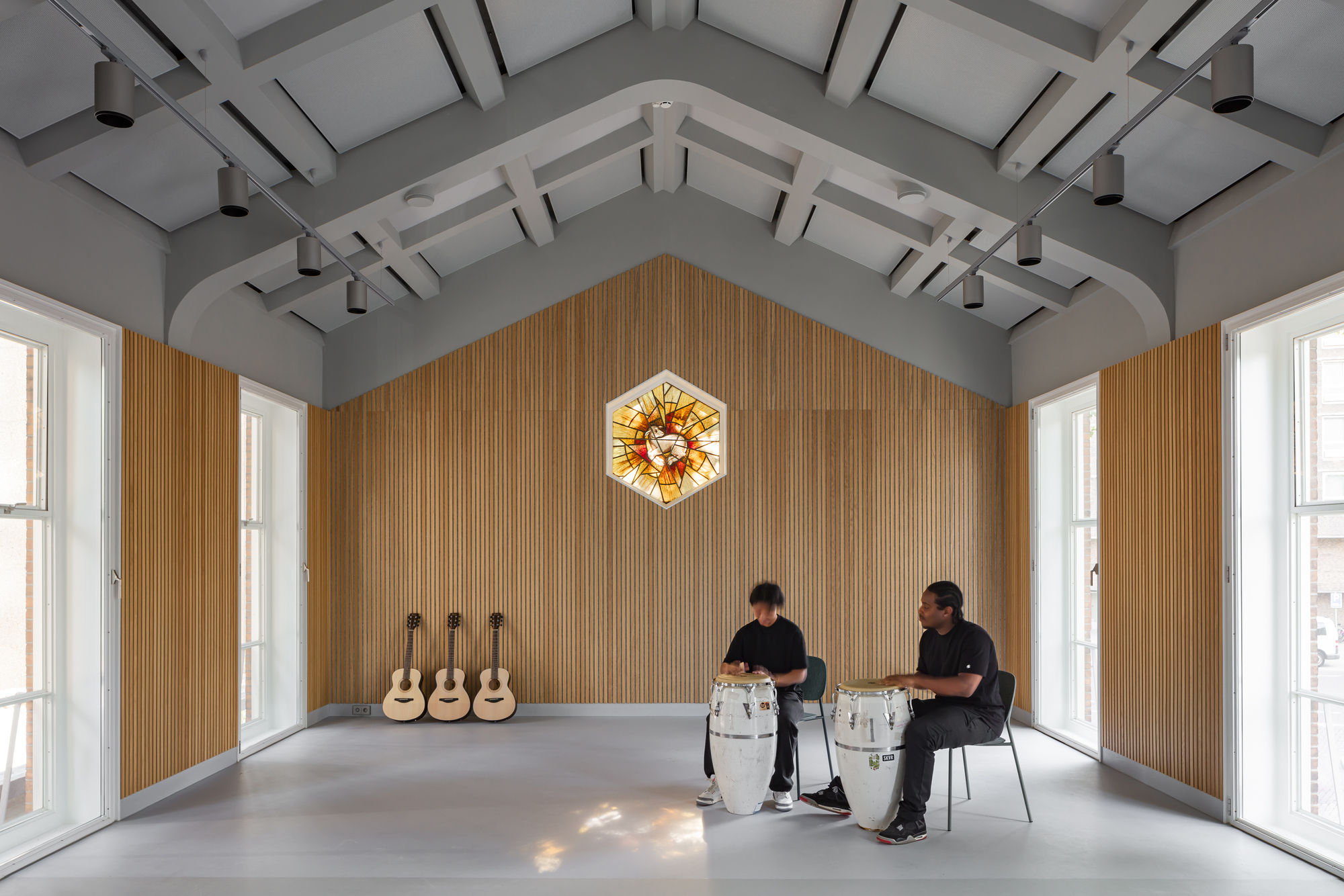 Vaulted rehearsal space with vertical timber wall cladding and two musicians playing hand drums