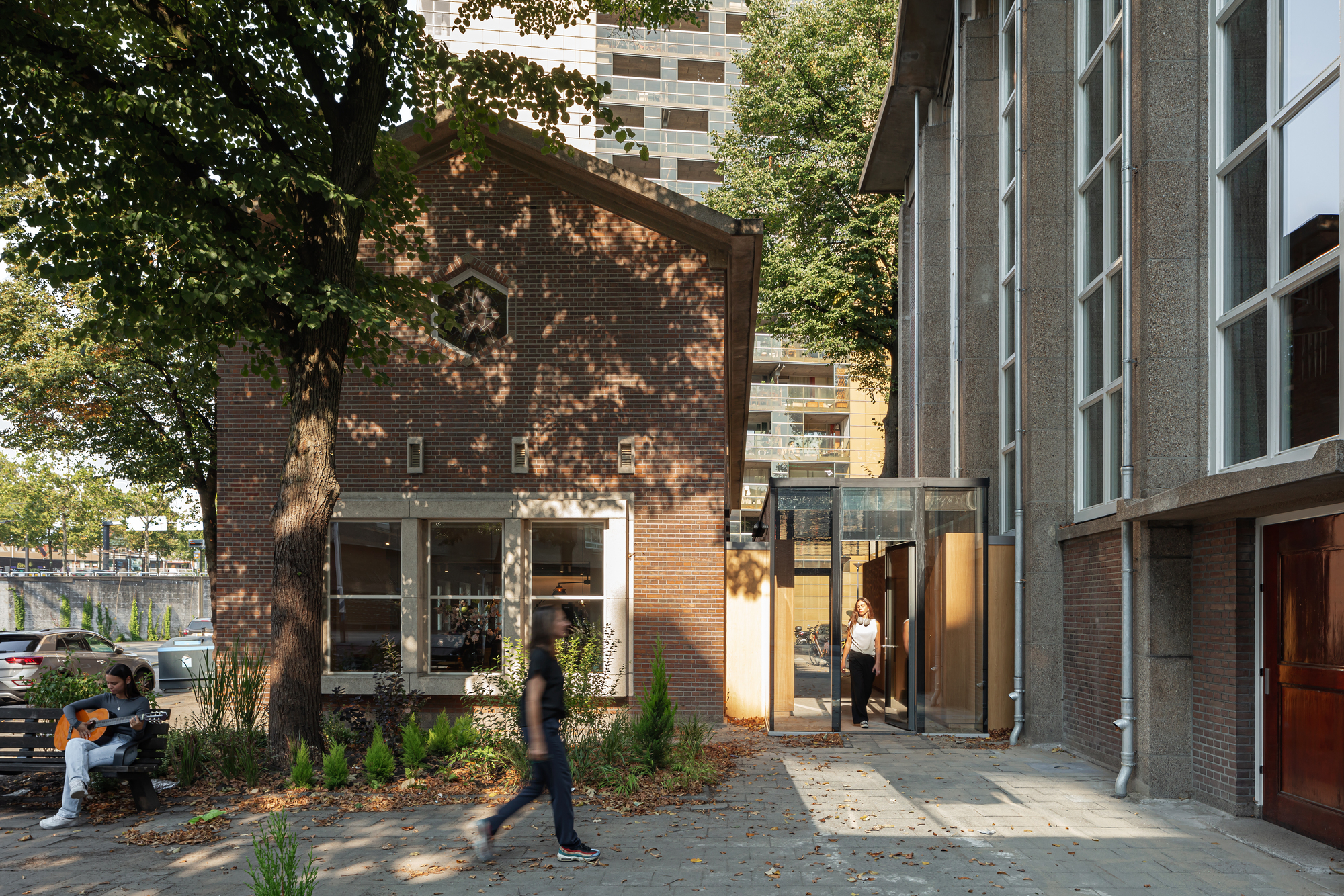 Brick gable end with ground-floor glazing viewed through dappled tree shade and planted beds