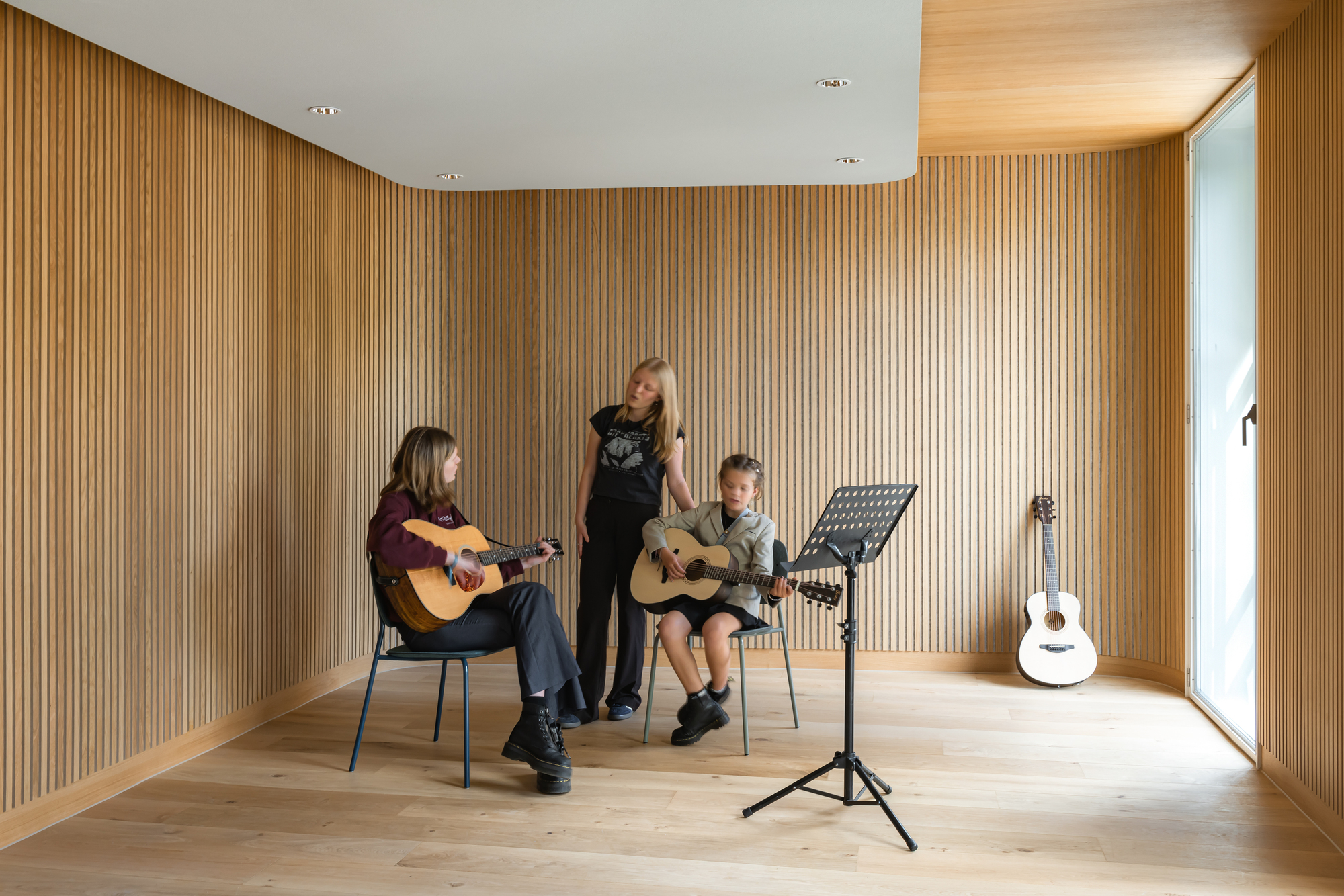 Music lesson in a timber-lined room with three people playing guitars beside a music stand