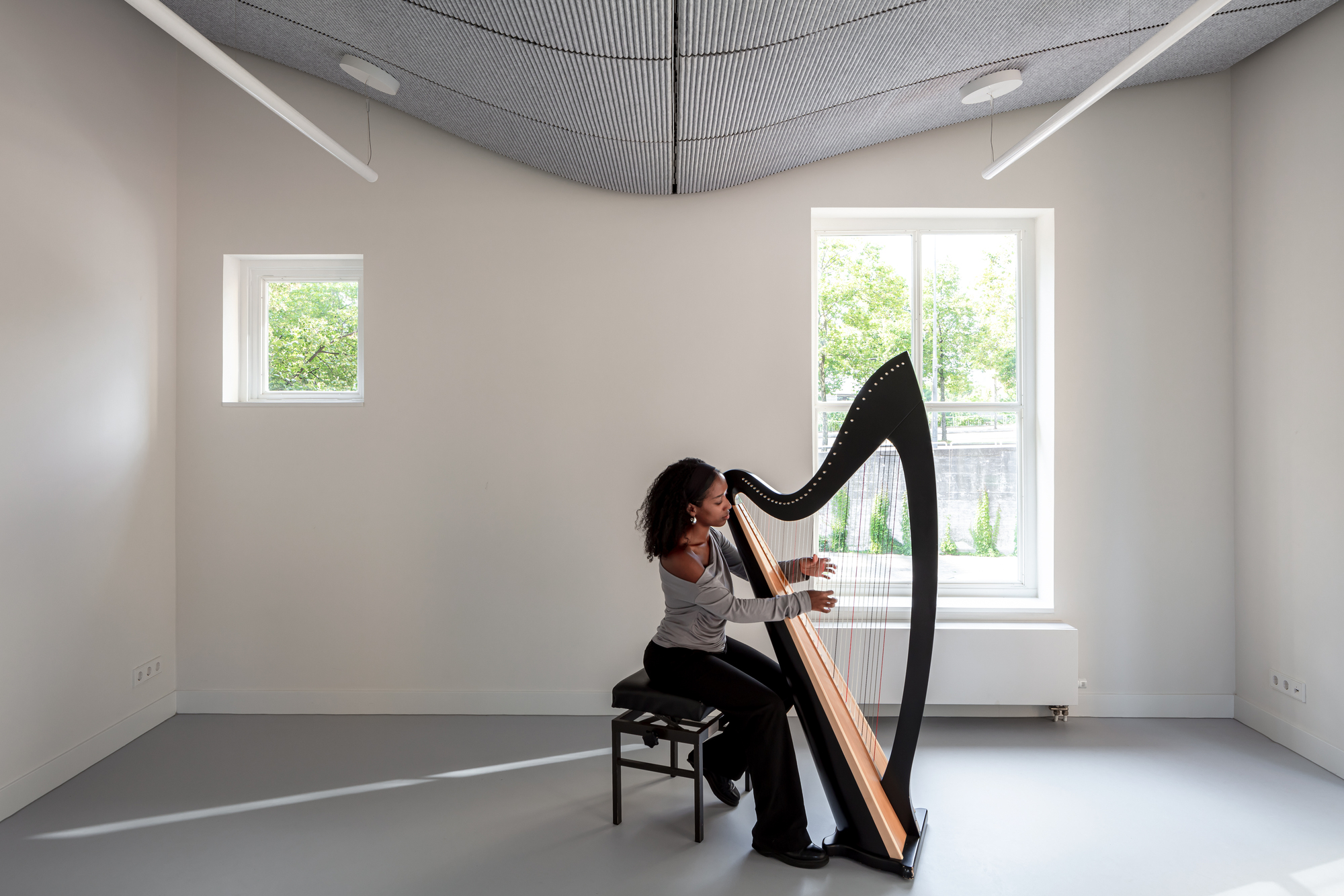 Practice room with curved acoustic ceiling panel, grey floor and musician playing harp by window