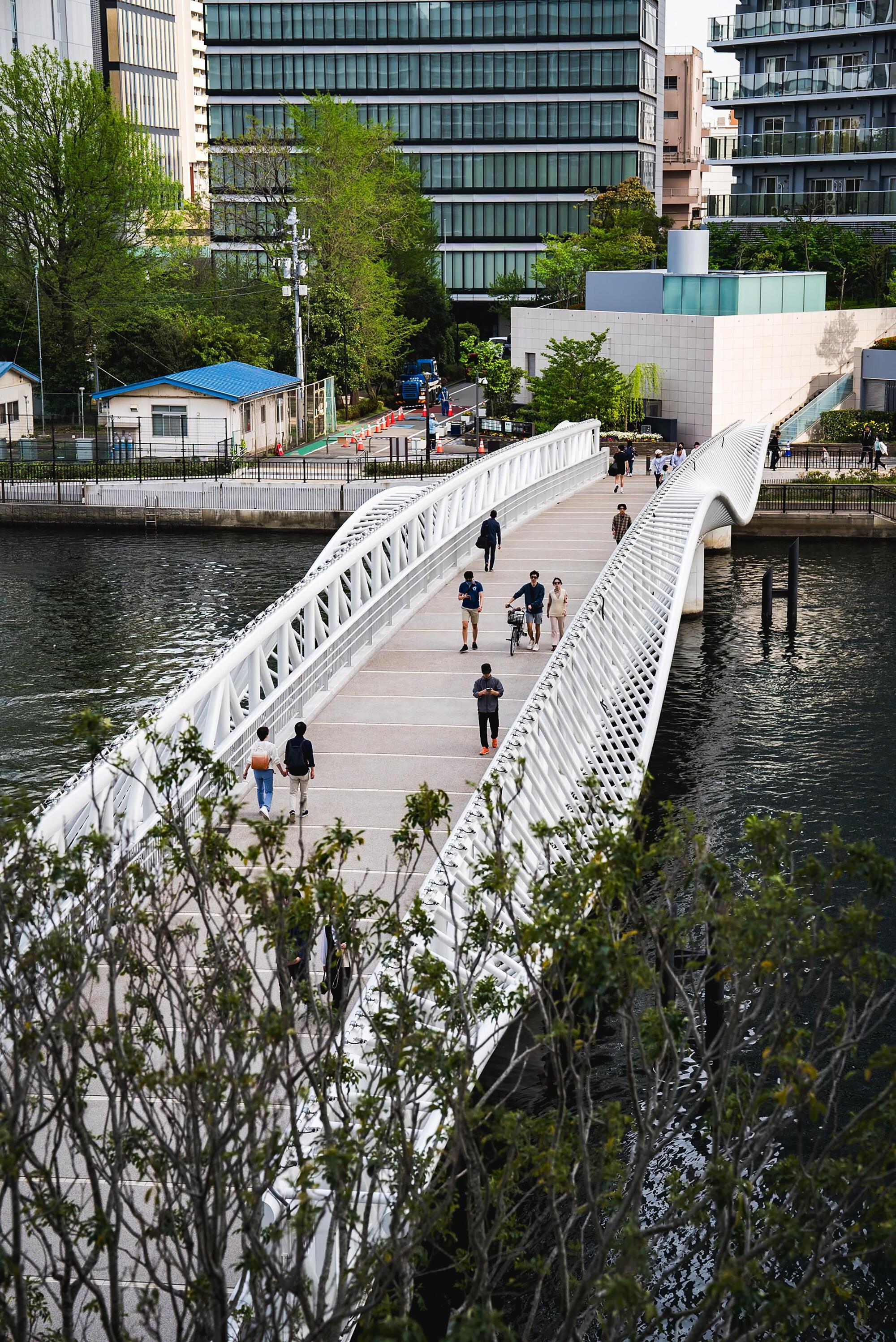 Pedestrian bridge deck with diagonal white lattice railings and cyclists crossing over still water