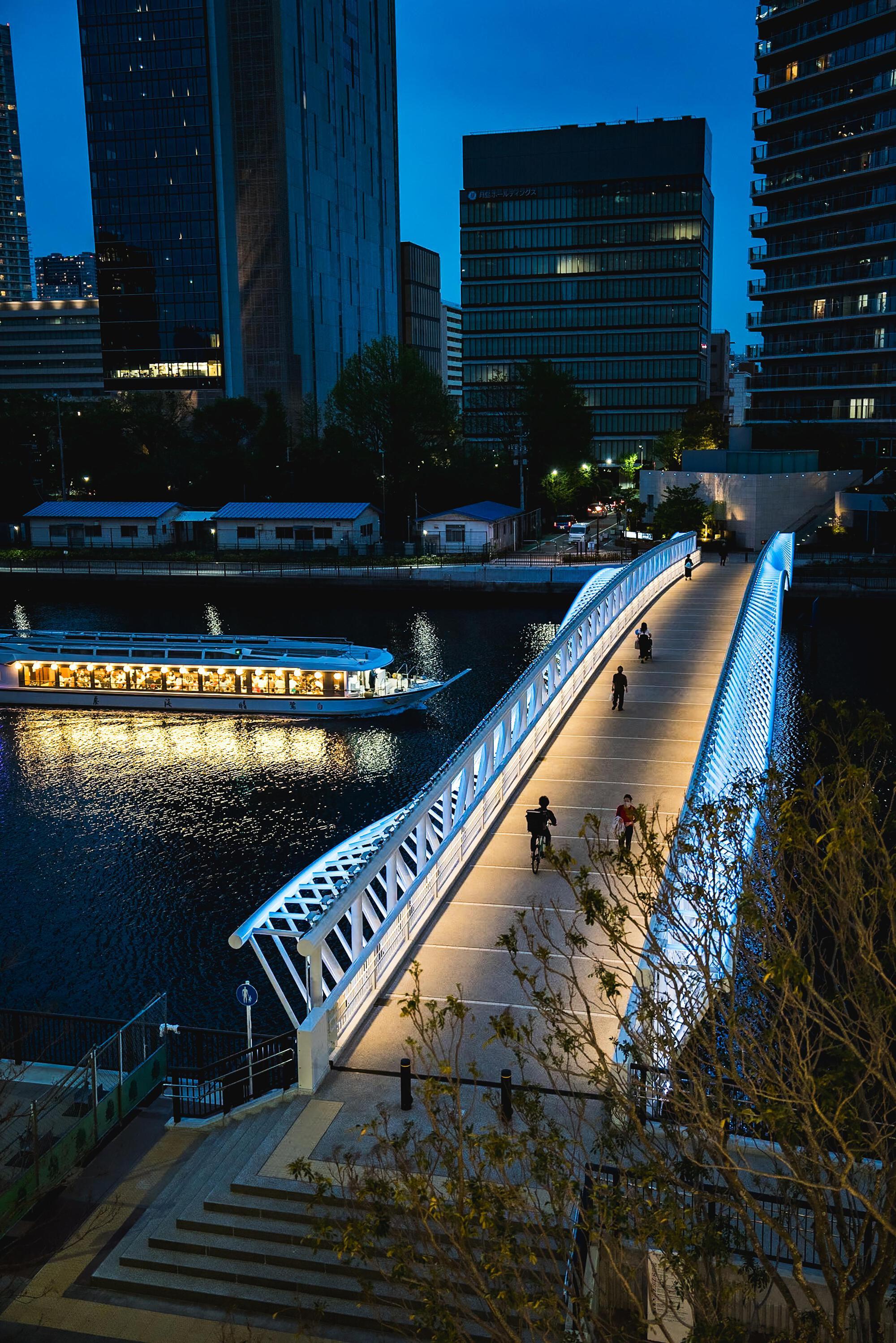Bridge illuminated at dusk with pedestrians crossing and a lit tourist boat passing below